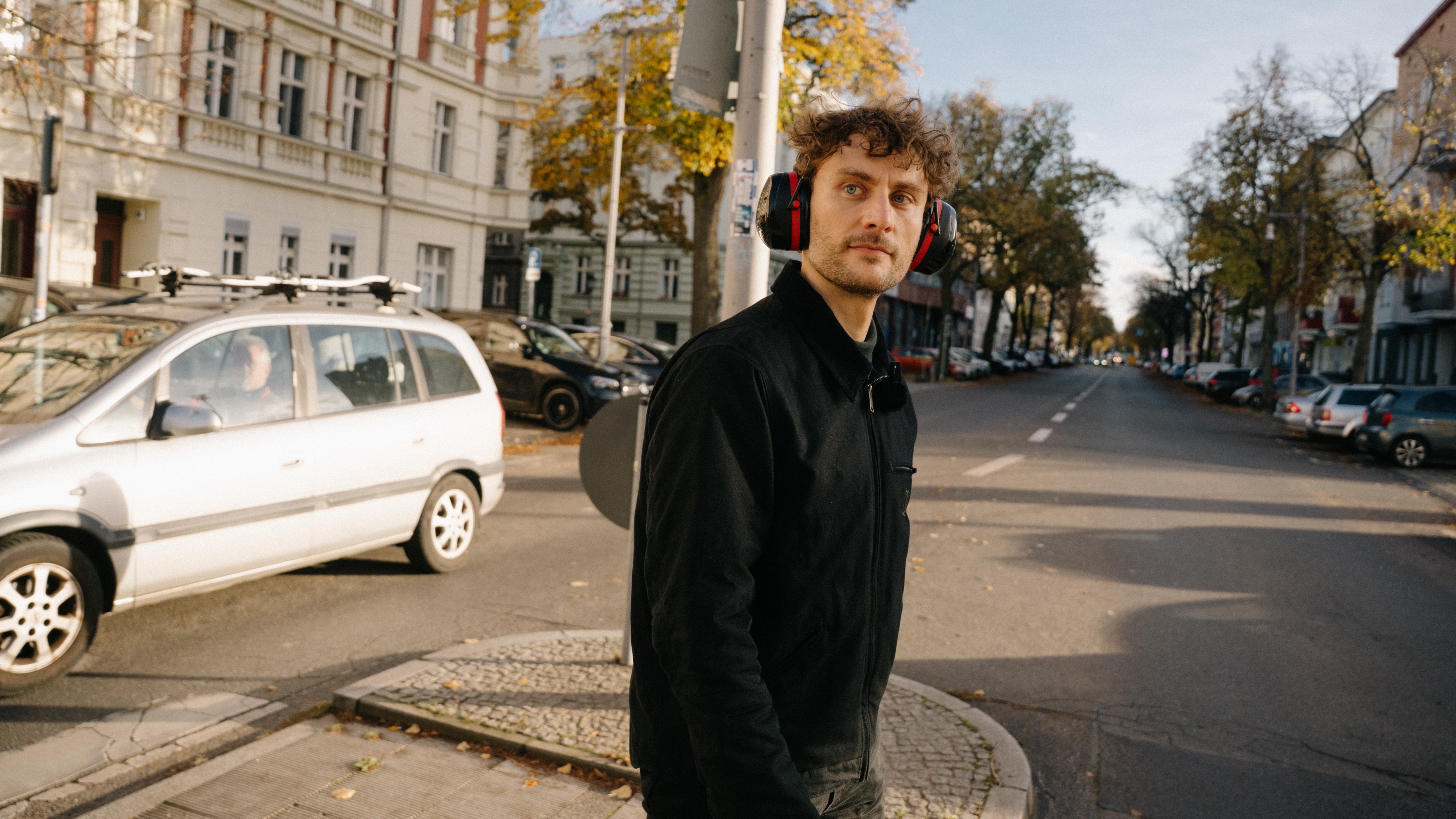 Ein Mann mit Gehörschutz steht auf einer Verkehrsinsel zwischen zwei Zebrastreifen einer befahrenen Straße in einer Stadt und blickt über seine Schulter. Im Hintergrund sind Häuser und am Straßenrand parkende Autos zu sehen.