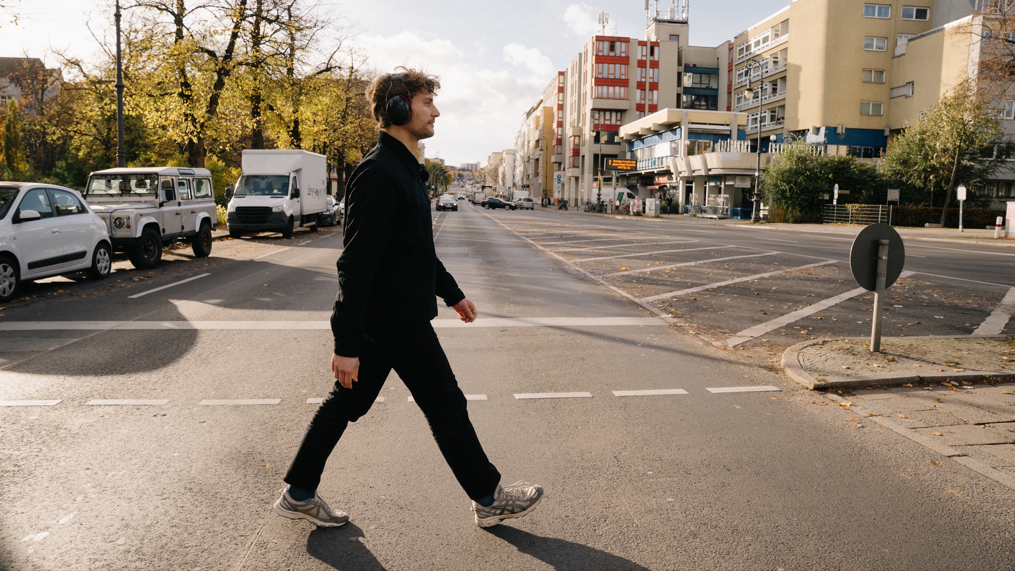 Seitenansicht eines gehenden Mannes mit Gehörschutz in dunkler Kleidung auf einem Fußgängerüberweg einer mehrspurigen Straße. Die Sonne scheint, am Straßenrand stehen Bäume mit Herbstlaub und moderne Gebäude.