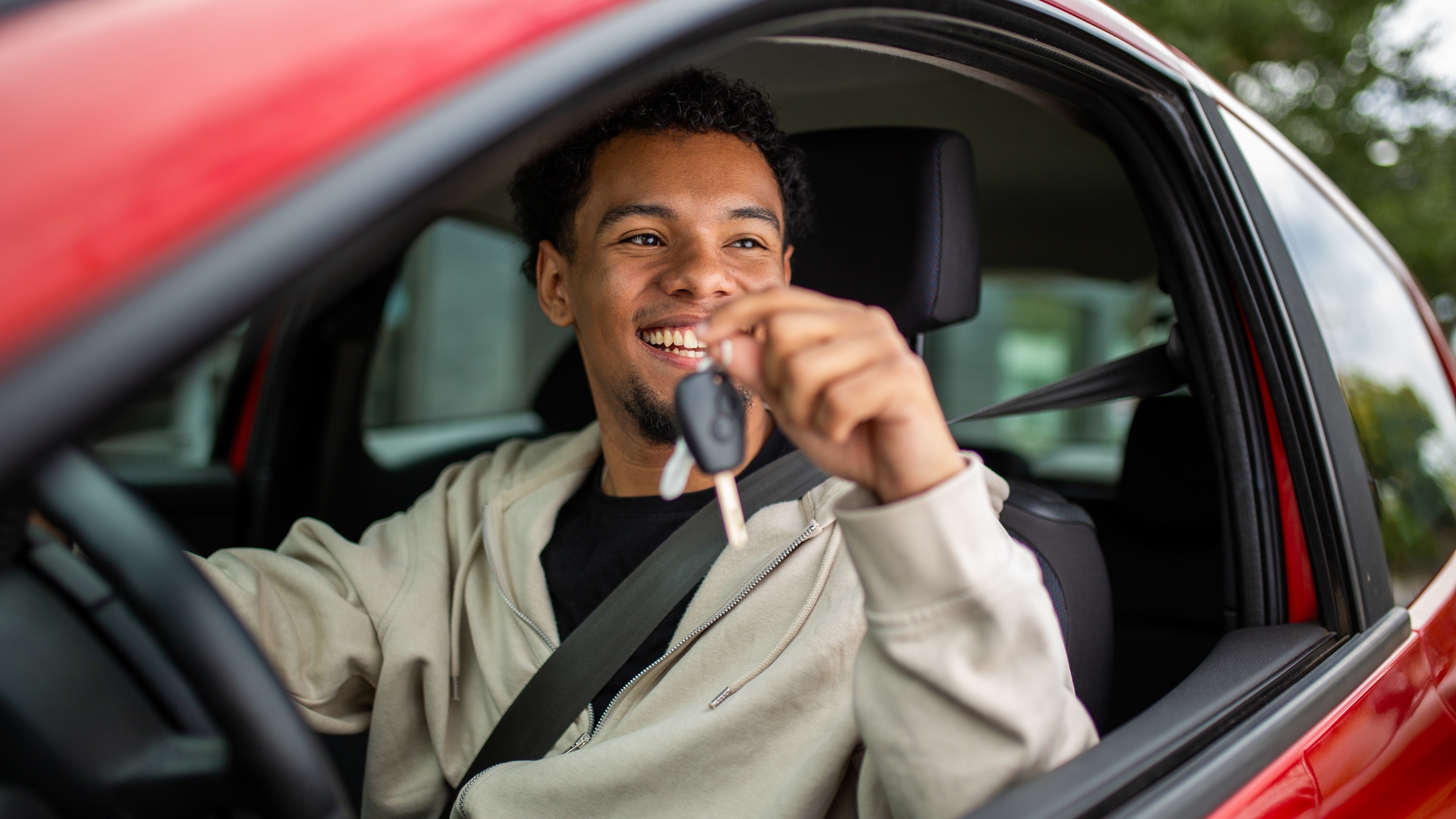 Ein junger Mensch mit kurzen schwarzen Haaren sitzt angeschnallt im Auto und hält einen Autoschlüssel in die Kamera. 