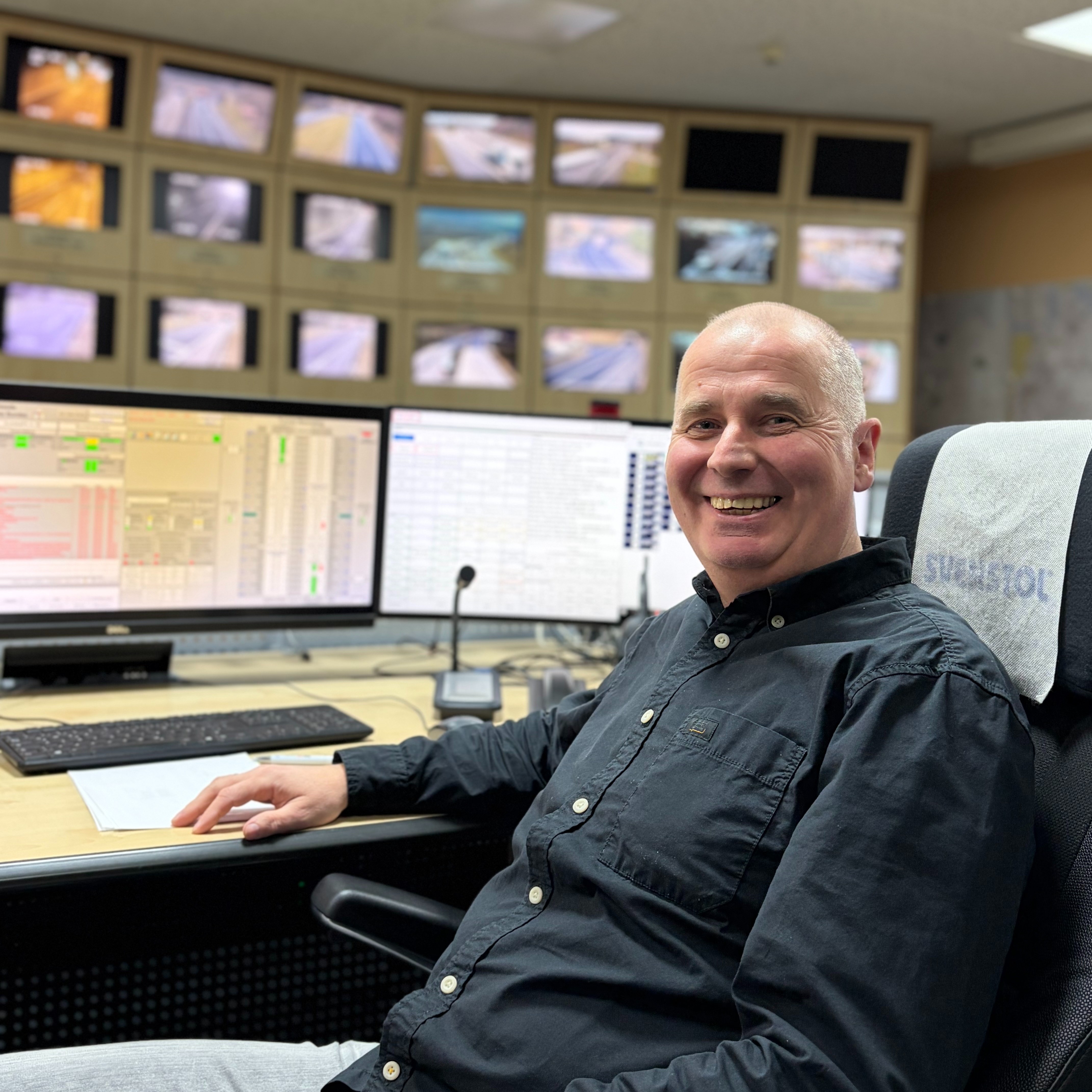 Porträt: Thomas Weyrich 1:1 Thomas Weyrich, ein lächelnder Mann, sitzt an einem Schreibtisch in einer Tunnelleitzentrale. Im Hintergrund sind zahlreiche Monitore zu sehen.