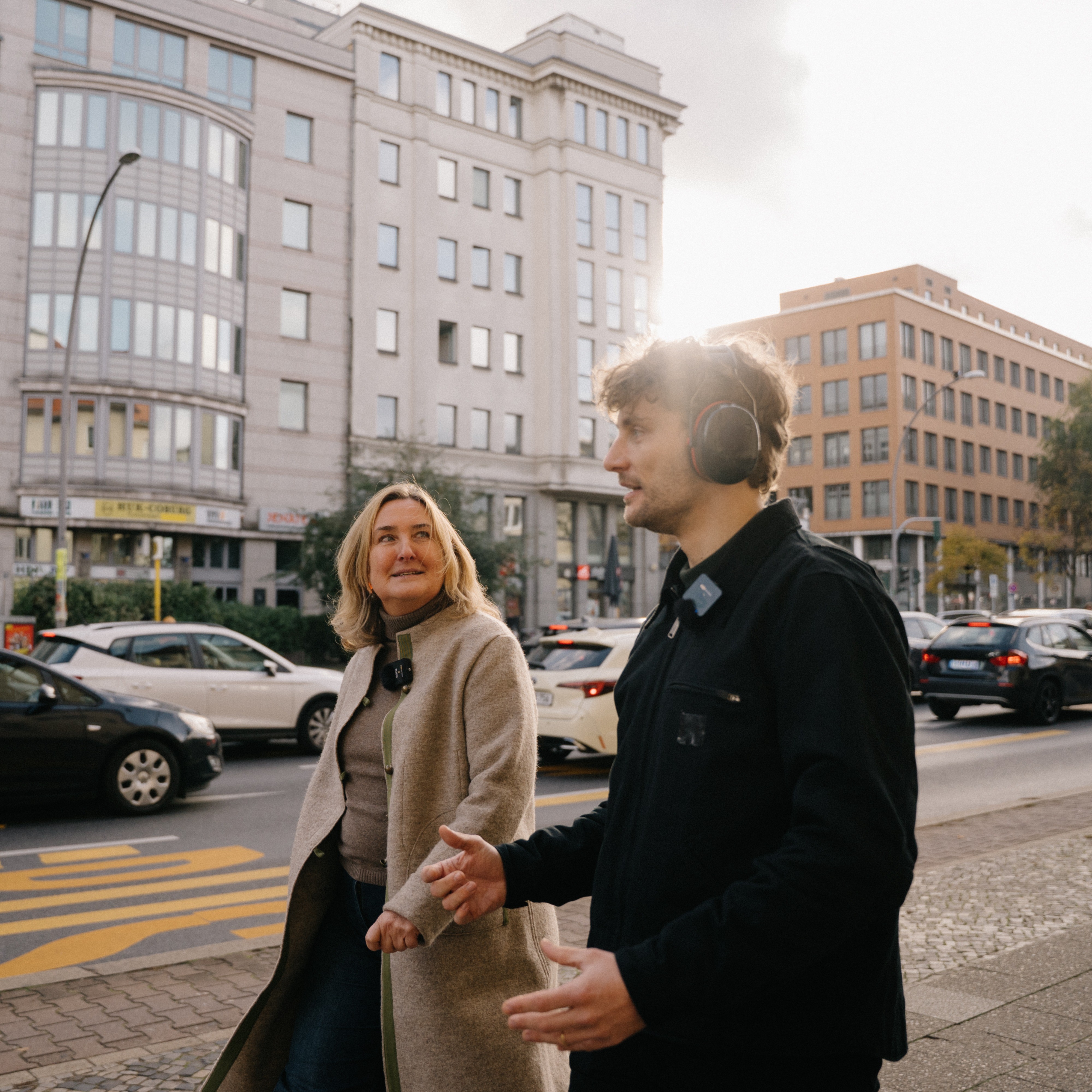 Seitenansicht eines Mannes mit Gehörschutz und einer Frau, die neben einer befahrenen Straße gehen. Im Gegenlicht der Sonne sind Gebäude und Autos im Stadtverkehr zu erkennen.