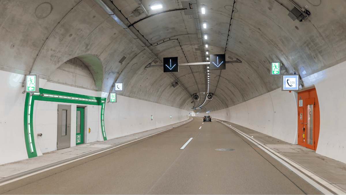 Innenansicht eines hell beleuchteten Straßentunnels mit einer Fahrspur. Auf der linken Seite ist ein grün gekennzeichneter Notausgang zu sehen. Auf der rechten Seite befindet sich eine orangefarbene Notrufstation mit einem Telefonsymbol darüber. Im Hintergrund fährt ein kleines Auto. 