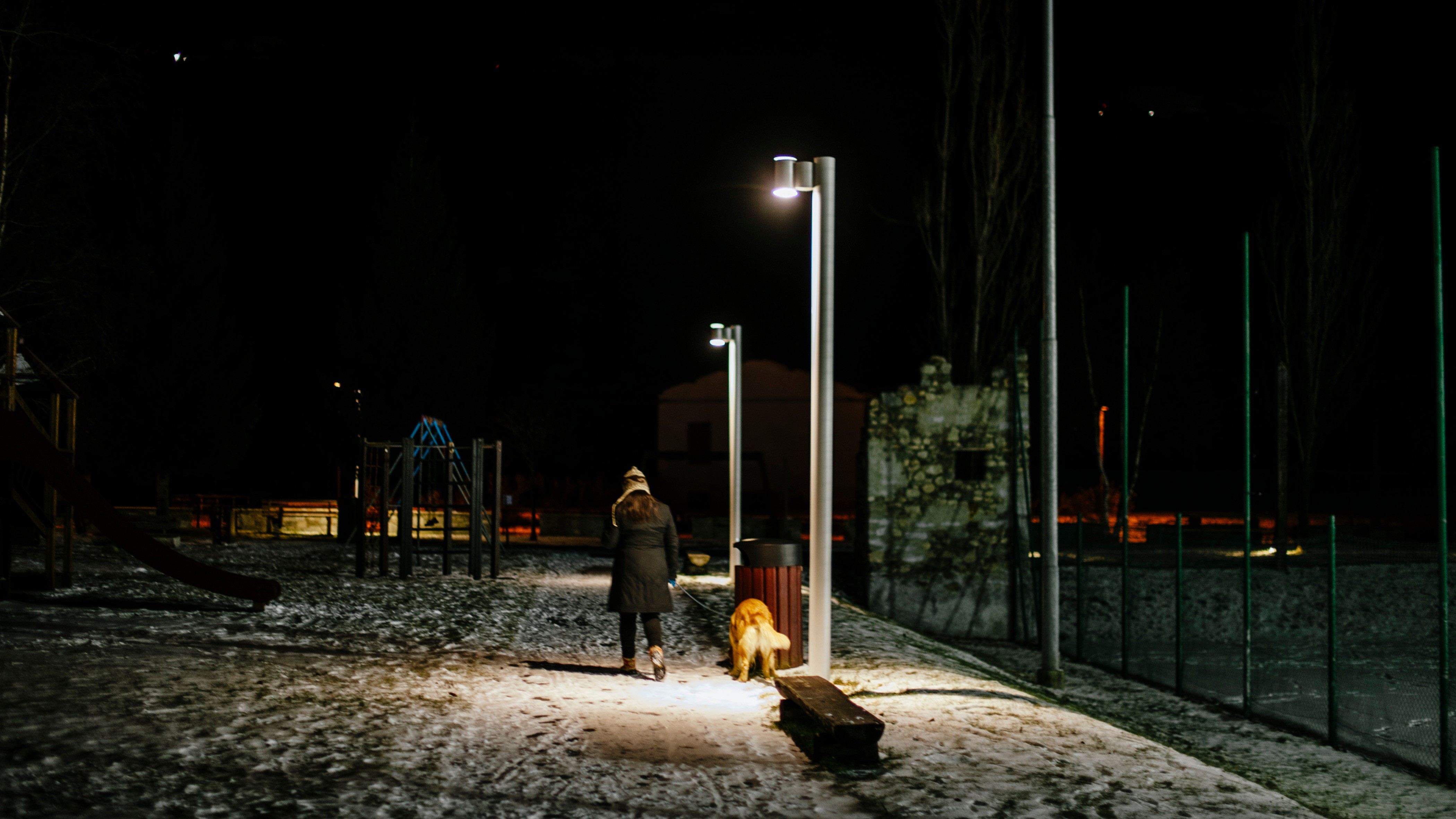 Gassi gehende Person bei Nacht (16:9) Zu sehen ist eine Frau, die gemeinsam mit ihrem Hund auf einem beleuchteten Gehweg in der Dunkelheit durch den Schnee spaziert.