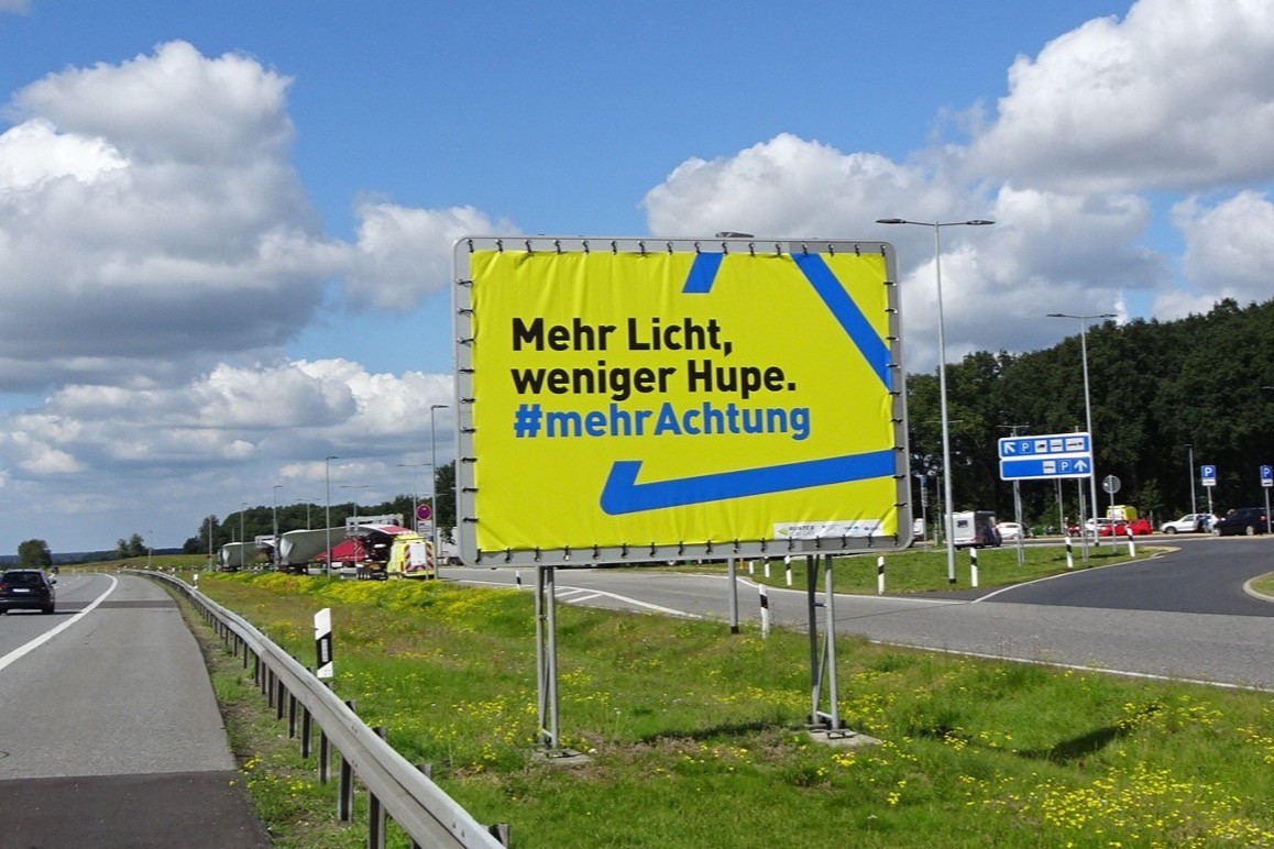 Am Rand einer Autobahn steht ein großer Plakataufsteller mit einem Autobahnplakat. Darauf ist Folgendes zu sehen: Vor einem blauen Dreieck auf gelbem Hintergrund, das an das Verkehrszeichen „Gefahrenstelle“ erinnert, steht der Text: „Mehr Licht, weniger Hupe. Mehr Achtung.“