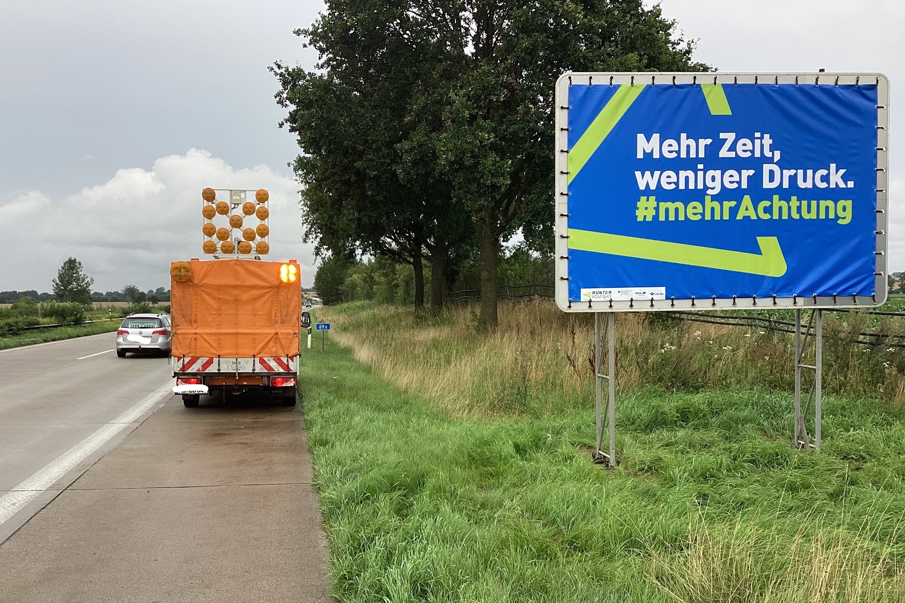 Neben einer Autobahn steht ein großer Plakataufsteller mit einem Autobahnplakat. Darauf ist Folgendes zu sehen: Vor einem hellgrünen Dreieck auf blauem Hintergrund, das an das Verkehrszeichen „Gefahrenstelle“ erinnert, steht der Text: „Mehr Zeit, weniger Druck. Mehr Achtung.“ Im Hintergrund sieht man einige Bäume und ein orangefarbenes Sicherungsfahrzeug der Autobahnmeisterei.