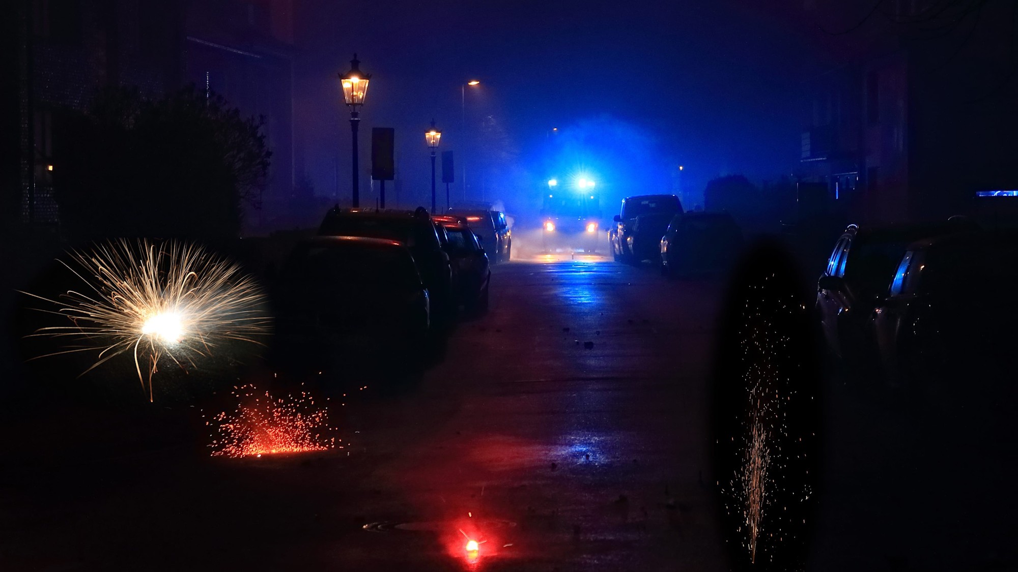 Bild Sicher durch die Silvesternacht 16:9 Ein Rettungsfahrzeug mit Blaulicht fährt durch eine dunkle Straße. Auf der Straße sind mehrere Feuerwerkskörper zu sehen.