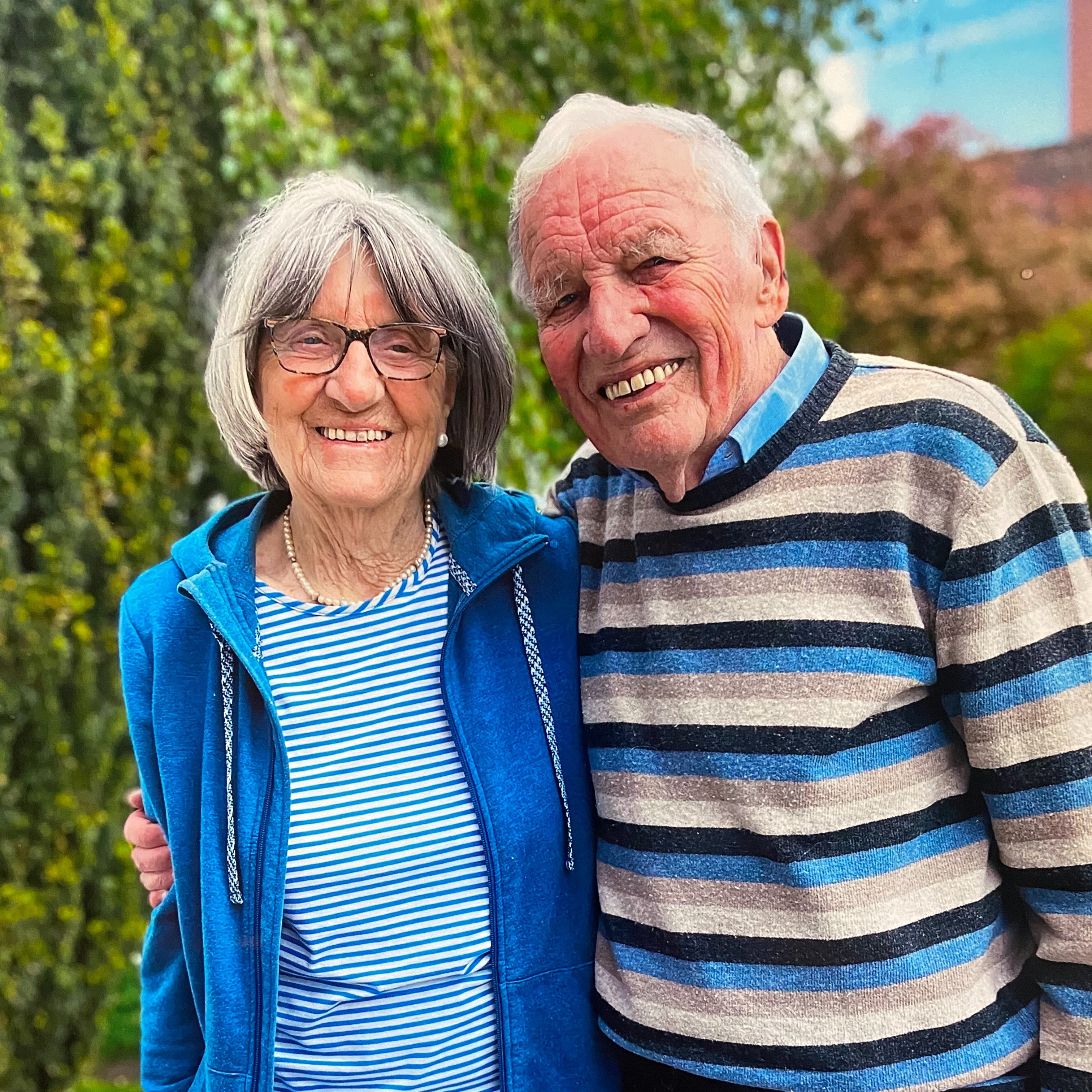 Bild mit 90 noch mobil Großeltern Eine Frau und ein Mann stehen Arm in Arm nebeneinander und blicken in die Kamera.