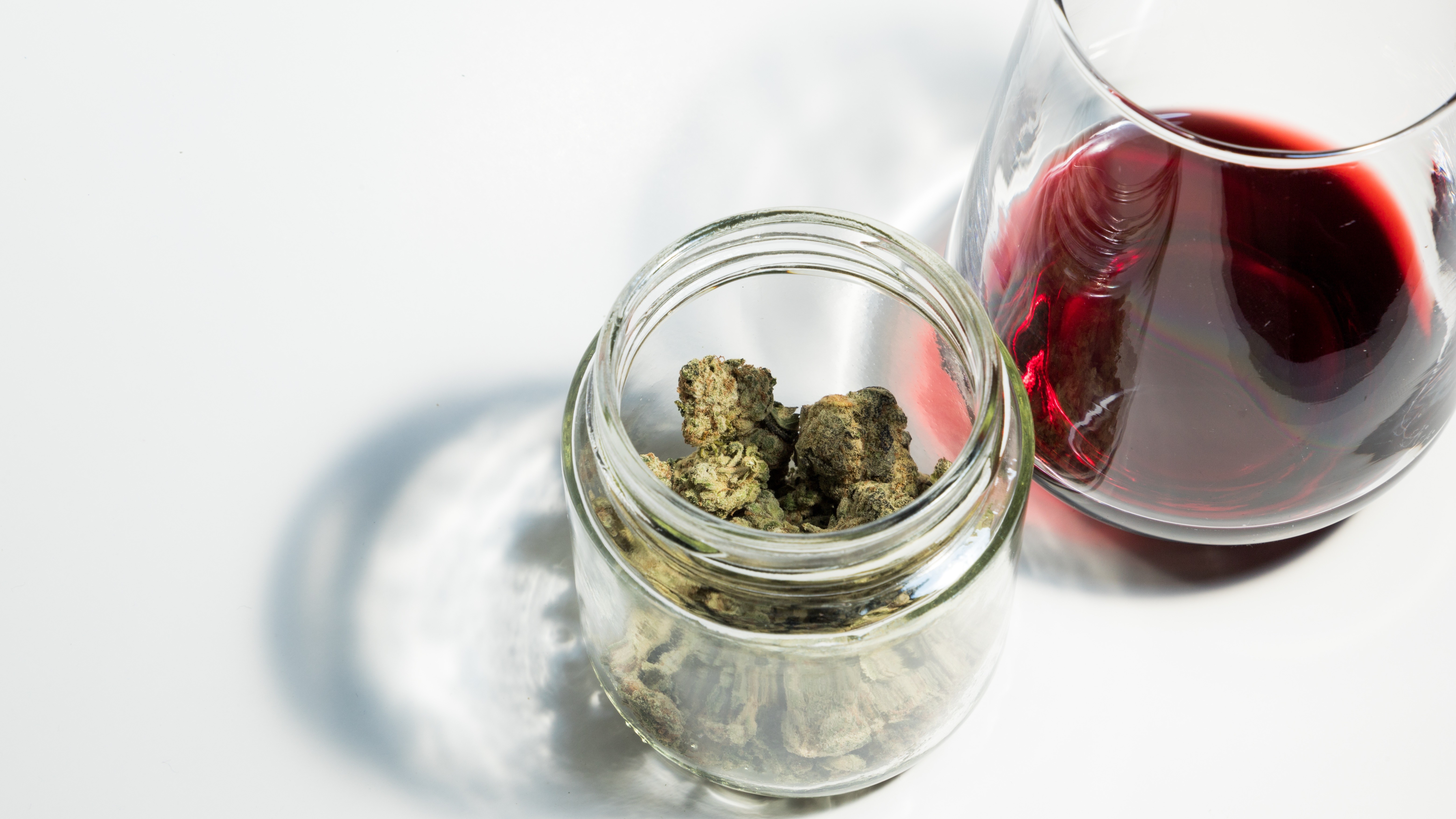 Weinglas und Cannabis 16:9 Das Bild zeigt ein kleines, durchsichtiges Glasgefäß, das mit getrockneten Cannabisblüten gefüllt ist. Rechts daneben steht ein Glas, das zur Hälfte mit Rotwein gefüllt ist. Beide Objekte befinden sich auf einer weißen Oberfläche.