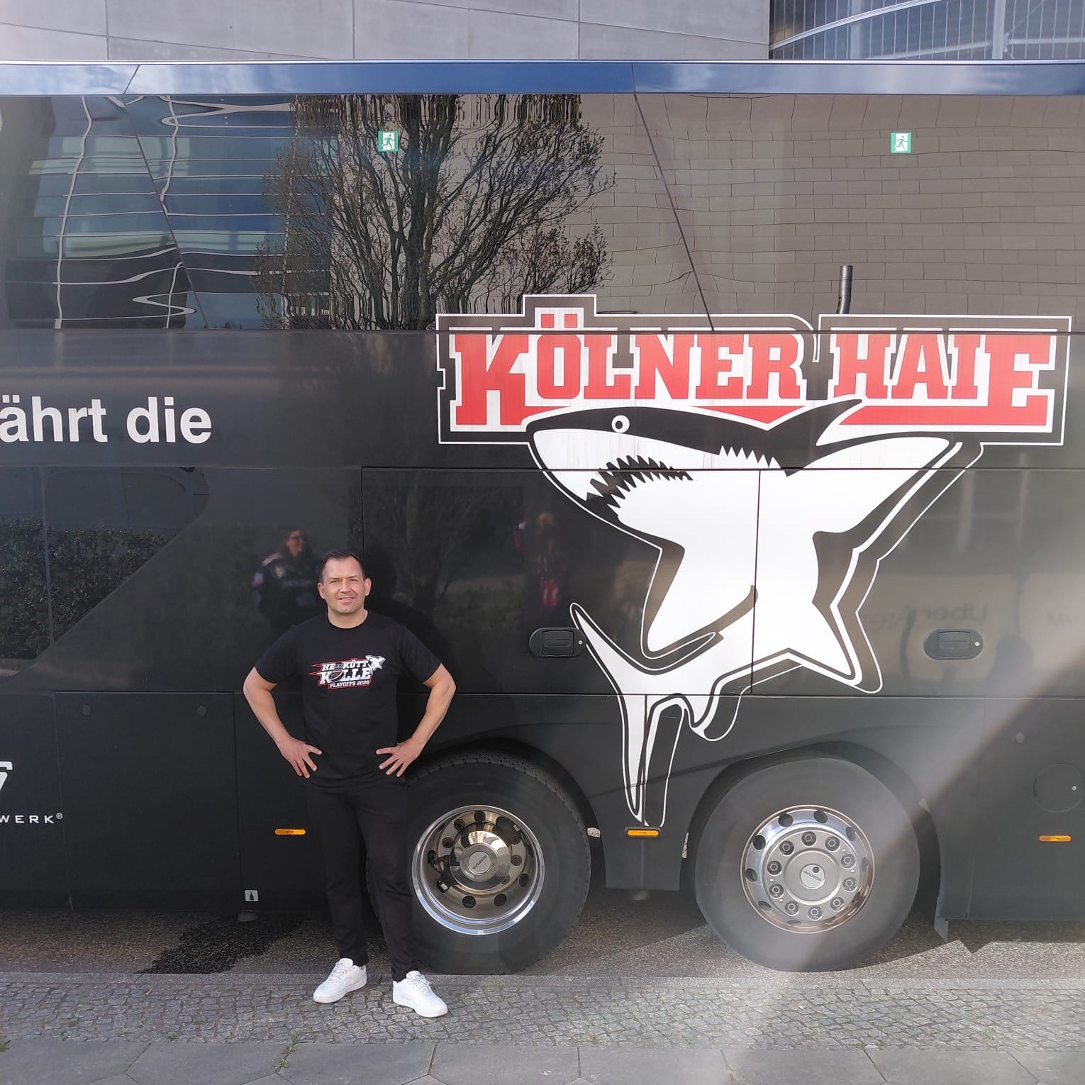 Busfahrer Enrico Bernig trägt einen roten „Kölner Haie“-Hoodie und zeigt das Peace-Zeichen. Im Hintergrund ist ein Bus zu sehen.