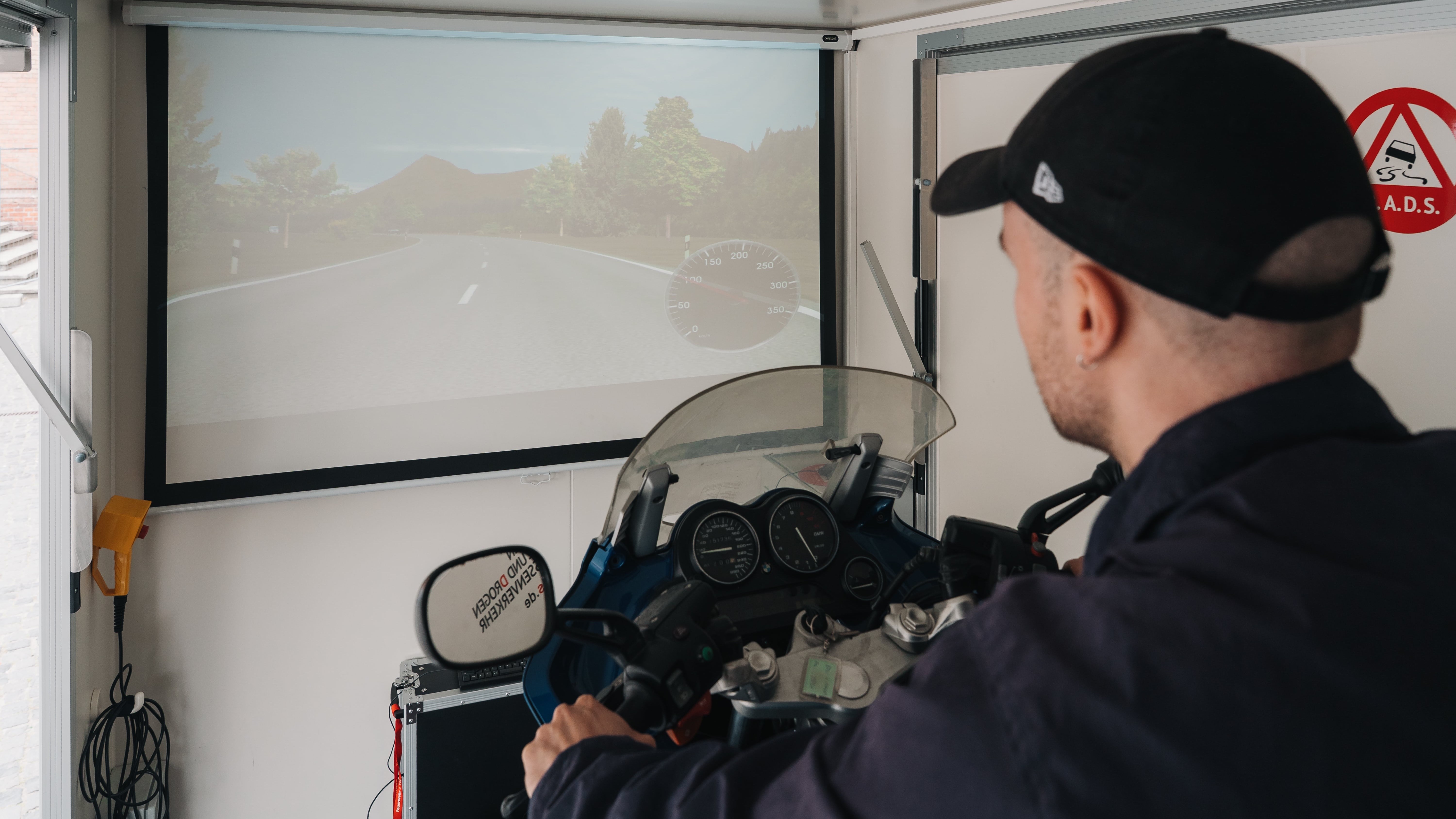 Eine Person testet einen Motorradsimulator in einem geschlossenen Raum. Auf dem Bildschirm erscheint eine virtuelle Straße mit Bäumen und Hügeln. Das digitale Cockpit mit Tacho und Spiegeln ist sichtbar. Ein Warnschild an der Wand verweist auf die Gefahren im Straßenverkehr.