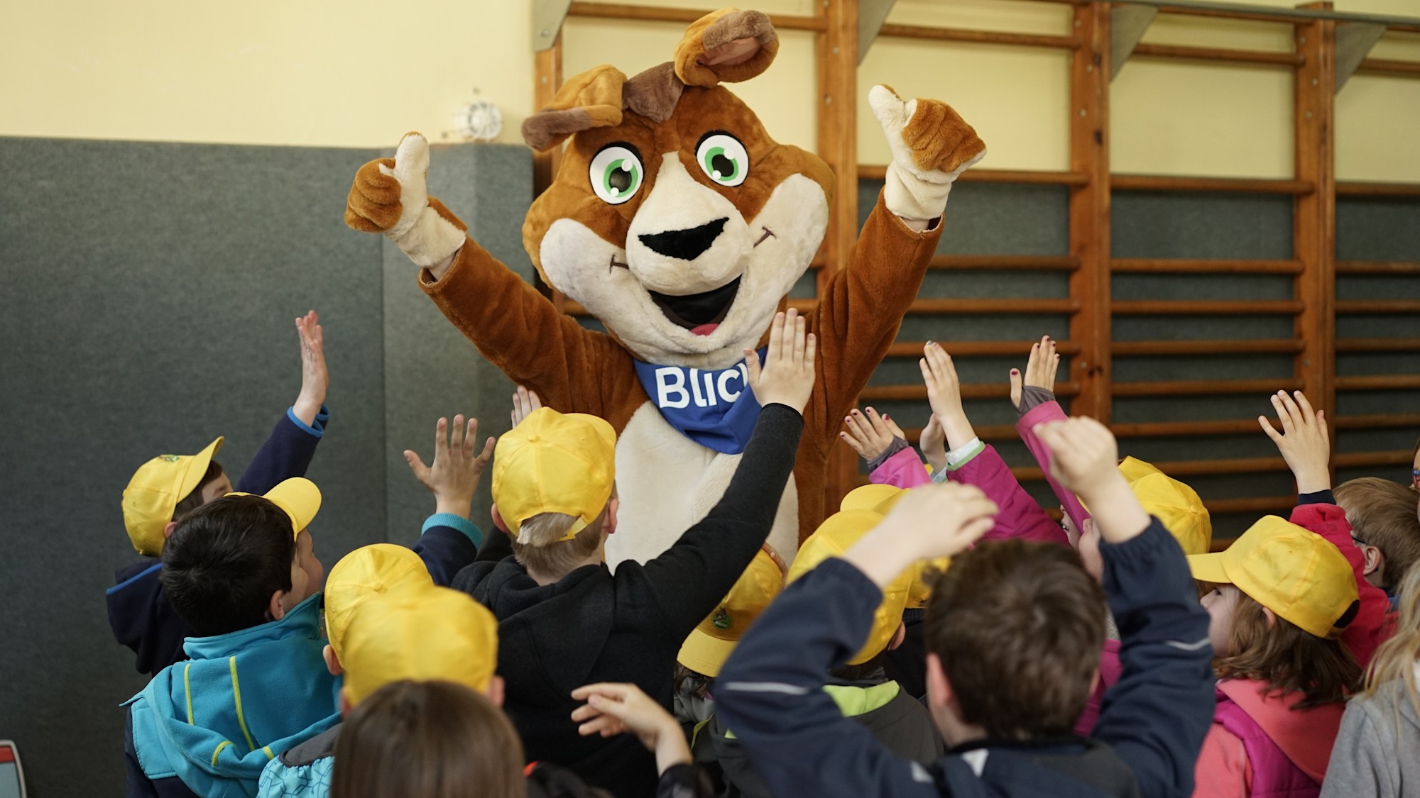 Mobilitaetsbildung leicht gemacht: Diese Spiele helfen beim Ueben Ein großes, braunes Känguru-Maskottchen mit einem blauen Halstuch, auf dem "Blicki" steht, steht in einem Raum mit einer Gruppe von Kindern, die gelbe Kappen tragen und ihre Hände in die Luft strecken. Blicki, das Känguru-Maskottchen, erklärt Kindern beim Verkehrssicherheits-Workshop die Verkehrsregeln.