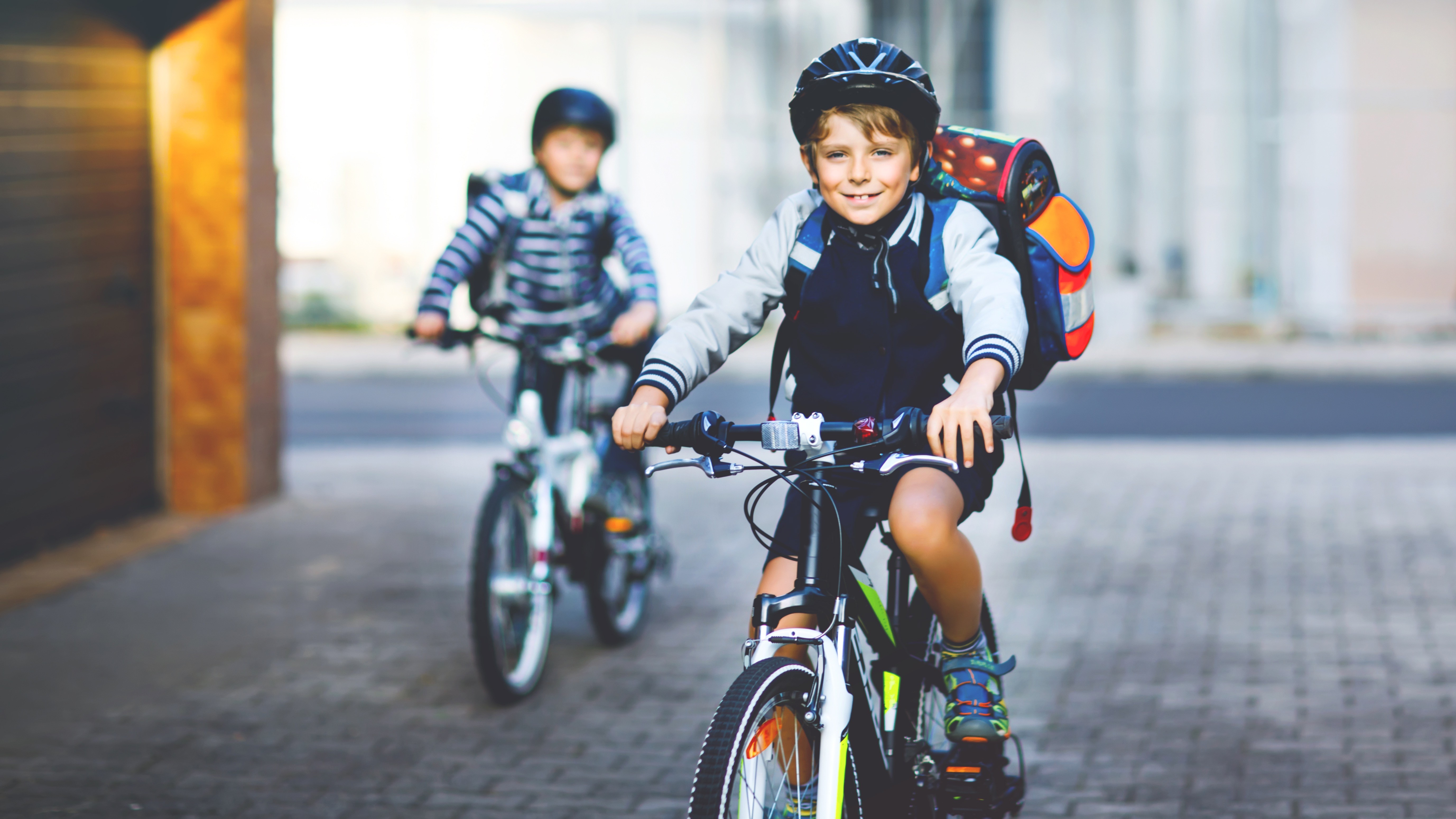 Zwei Grundschulkinder mit Helmen und Schulranzen fahren mit dem Fahrrad sicher zur Schule.
