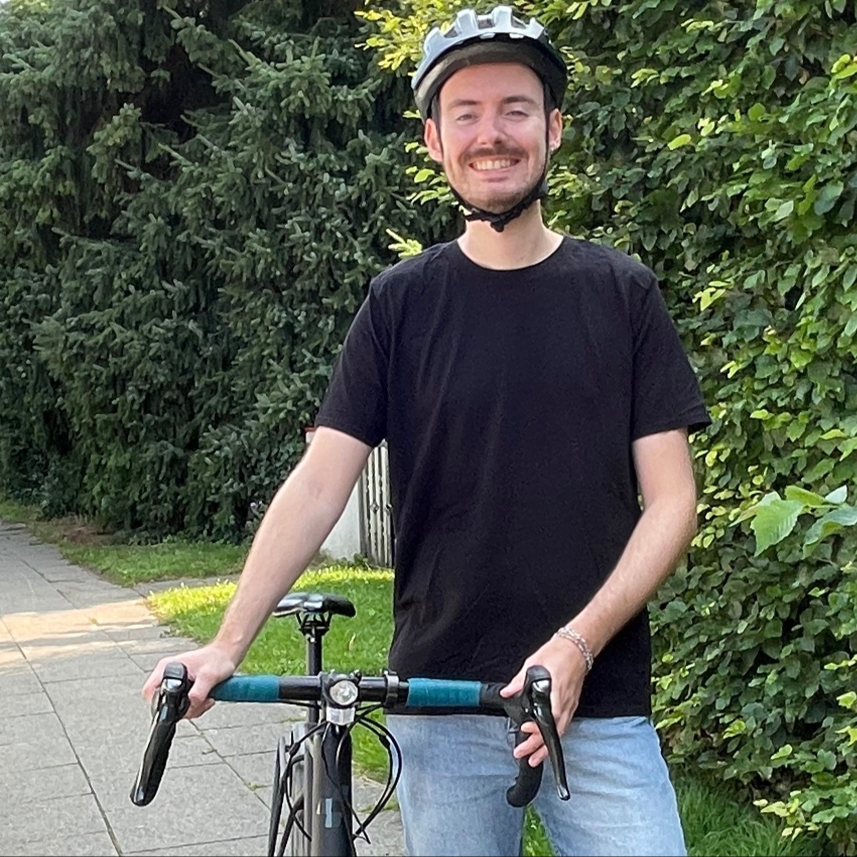 Ein junger Mann steht neben seinem Fahrrad und lacht freundlich. Hinter ihm sieht man Bäume und hohe Büsche sowie eine Straße mit parkenden Autos. 