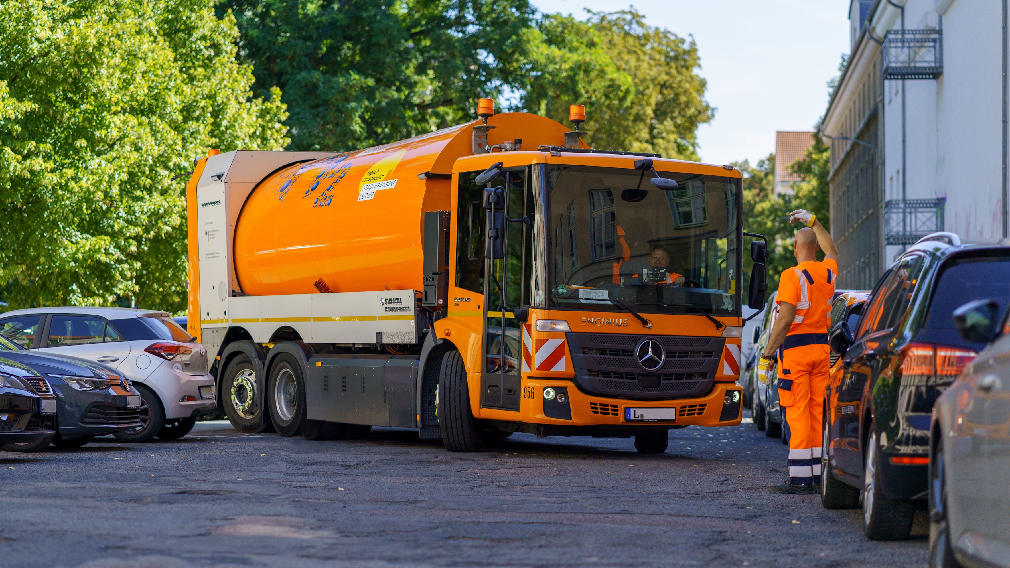 Ein Müllwagen mit Einweiser in enger Straße zwischen parkenden Autos. 