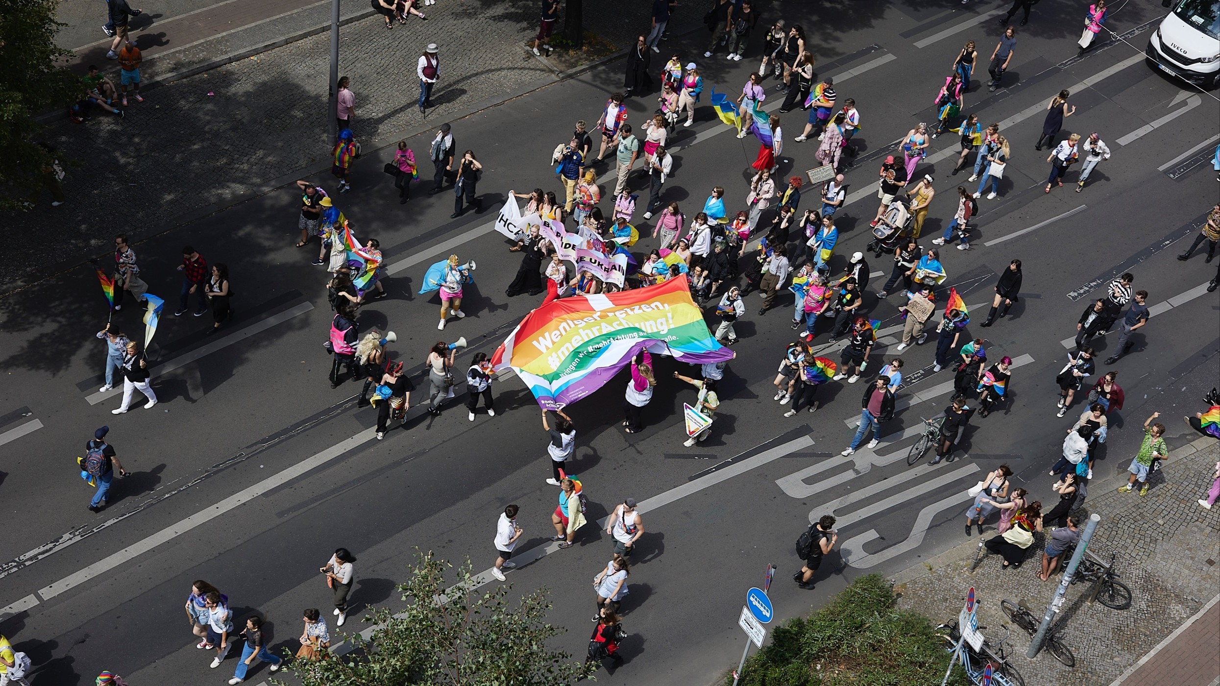 Teilnehmende einer Parade laufen eine Straße entlang. Sie halten ein Banner mit der Aufschrift: „Weniger hetzen, mehr Achtung!“