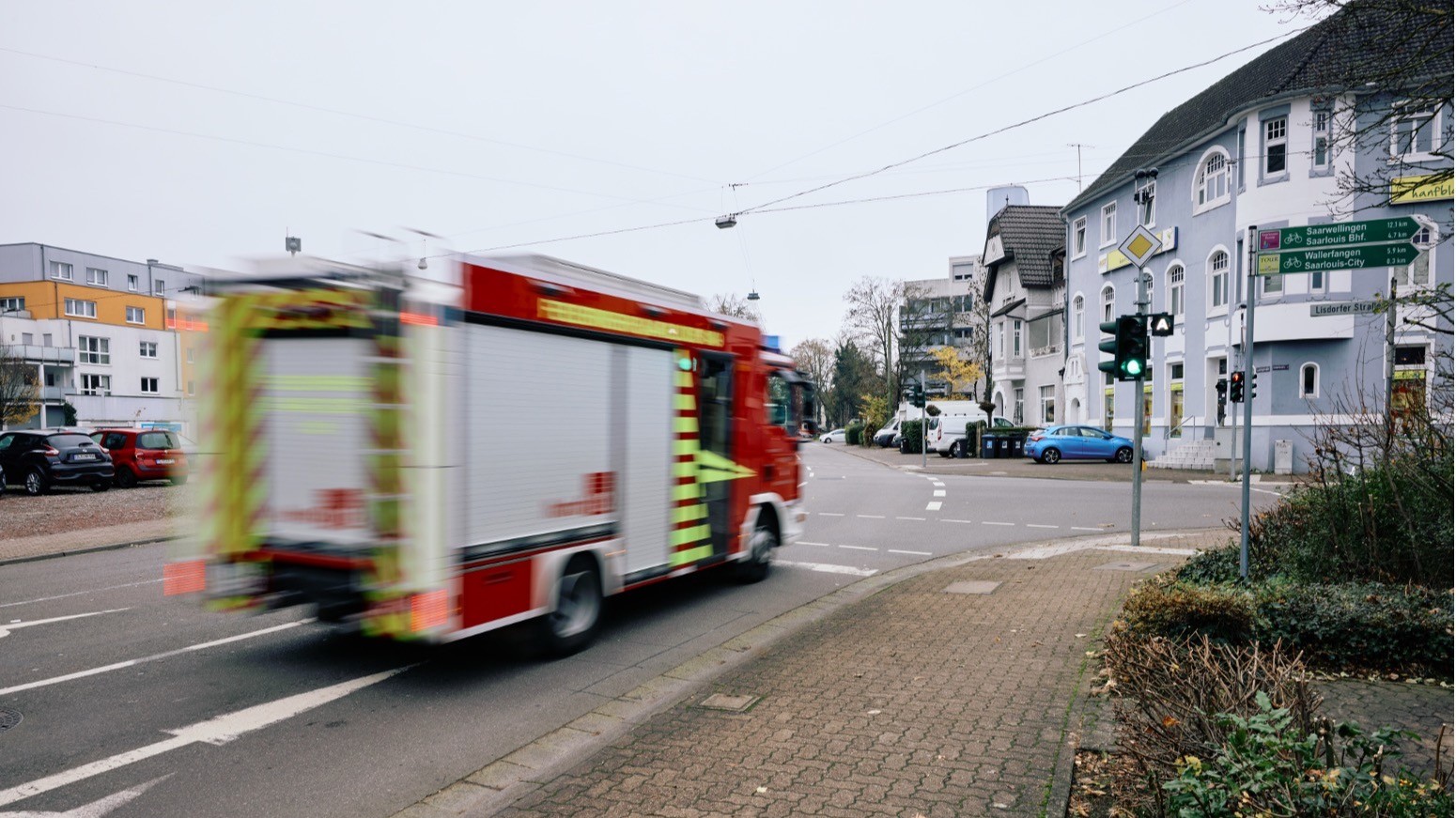 Ein Feuerwehrauto fährt über eine Kreuzung. Das Fahrzeug ist leicht verschwommen. Im Hintergrund sind Hausfassaden und parkende Autos zu sehen. 