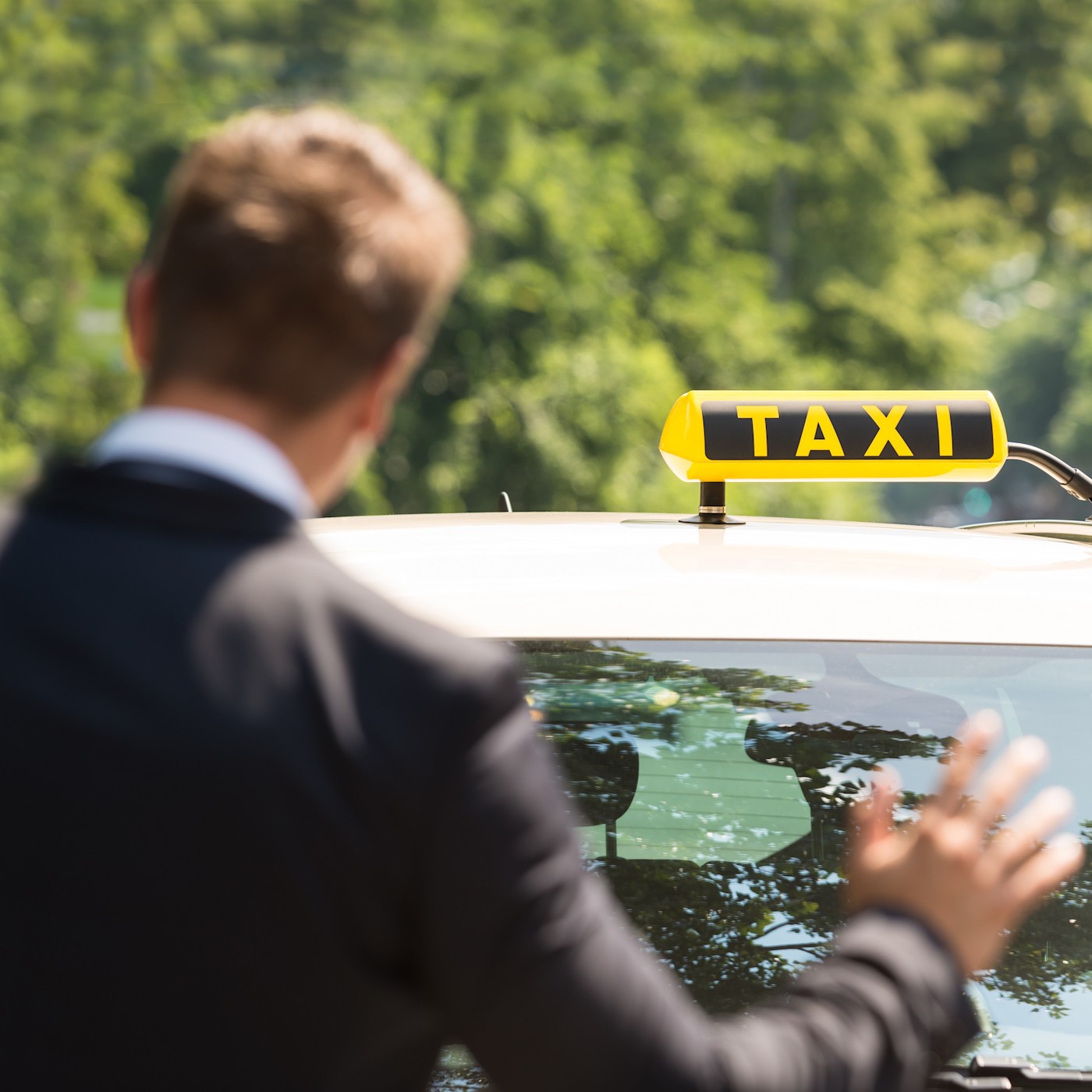 Bild Im Taxi (1:1) Ein Mann in einem Anzug steht mit dem Rücken zur Kamera und winkt ein Taxi heran. Das Taxi hat ein gelbes Schild auf dem Dach mit der Aufschrift „Taxi“. Der Hintergrund ist verschwommen und zeigt Bäume und eine Straße.