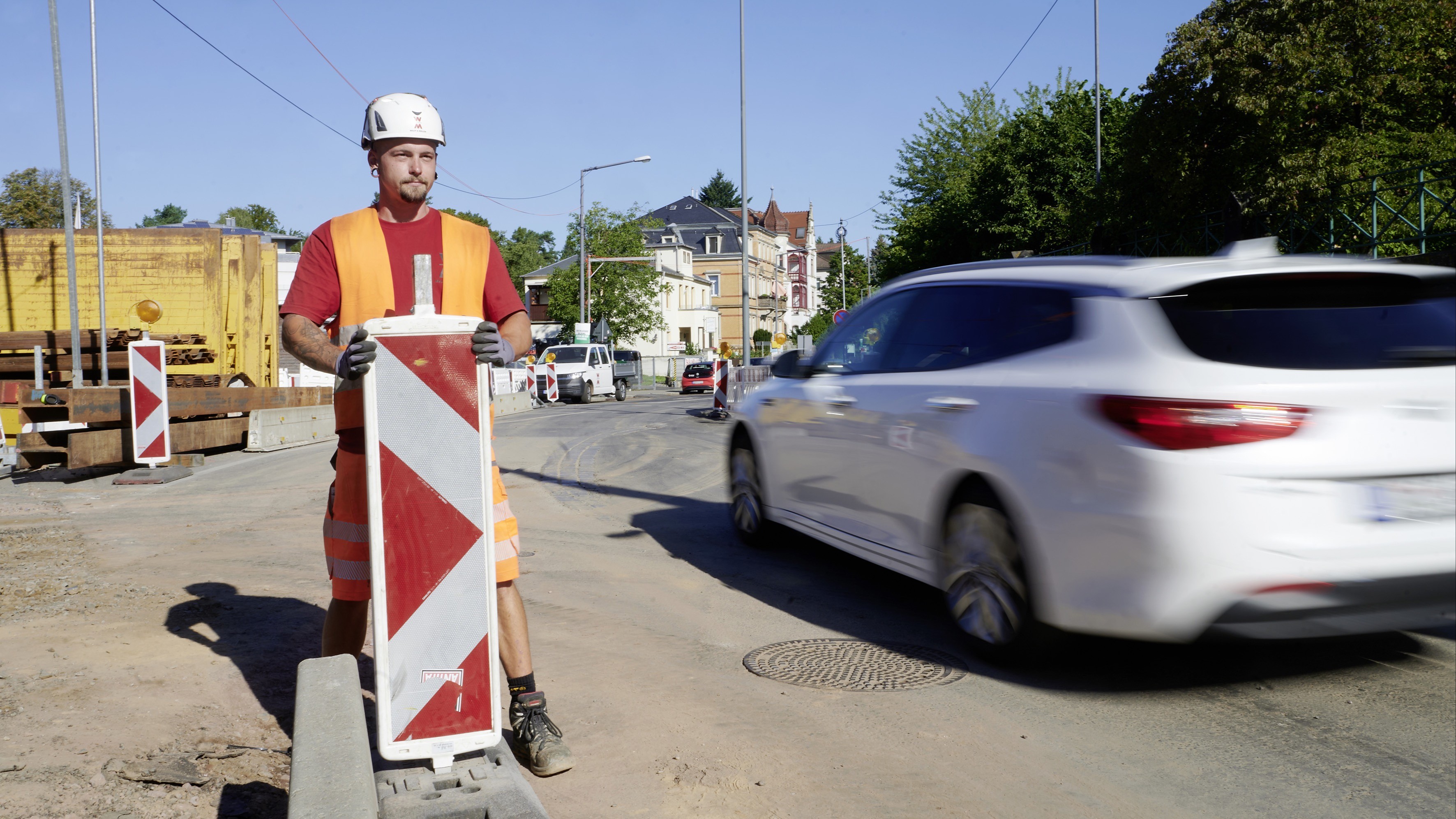 Ein Straßenarbeiter in orangefarbener Warnweste und Helm steht neben einer rot-weiß gestreiften Leitbake auf einer Baustelle. Im Hintergrund fährt ein weißes Auto vorbei.
