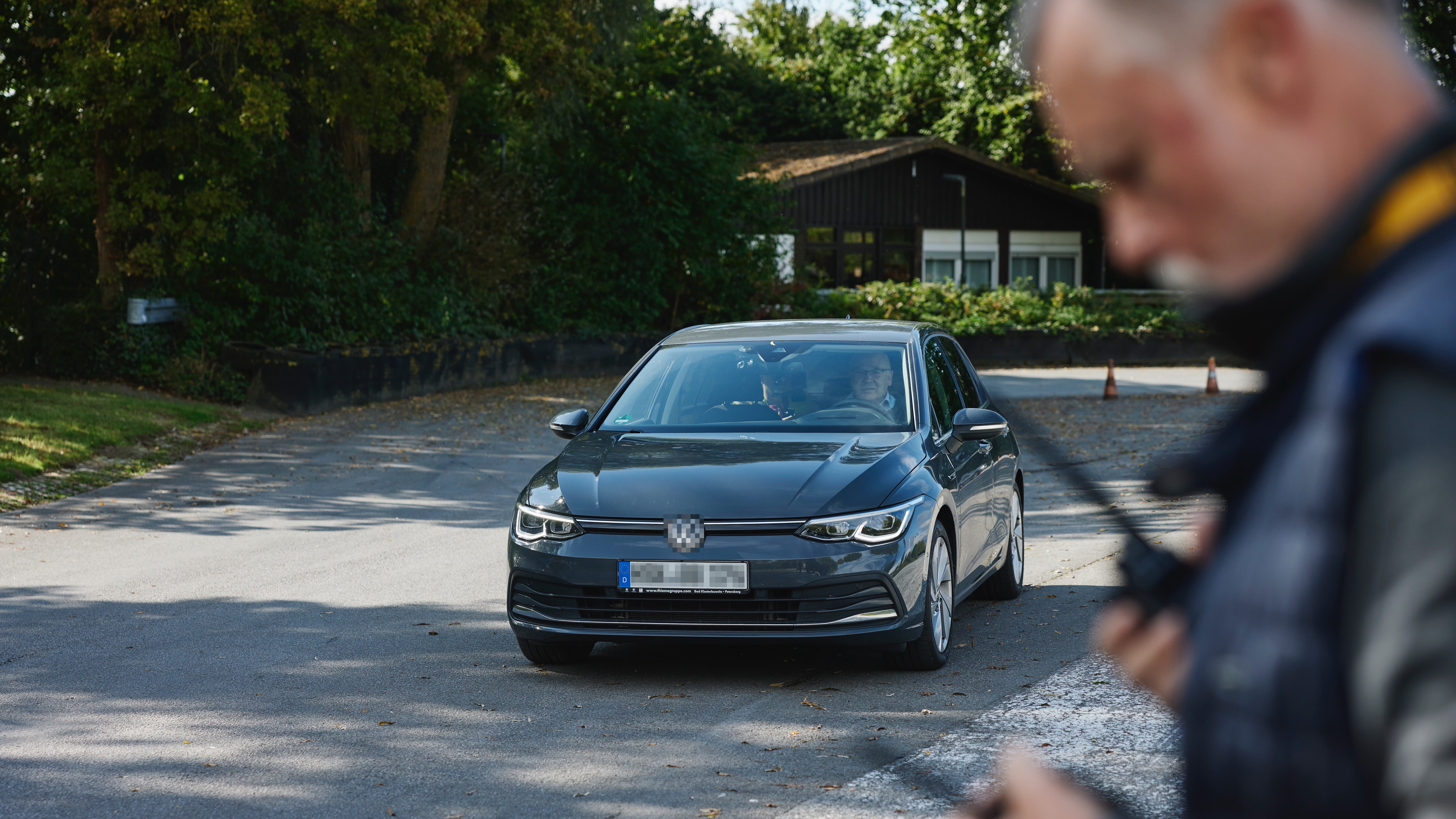 Fahrsicherheitstraining Strasse und Funkgeräte Ein graues Auto steht auf einer Straße, umgeben von Bäumen und einem Gebäude im Hintergrund. Im Vordergrund hält eine Person zwei Funkgeräte.