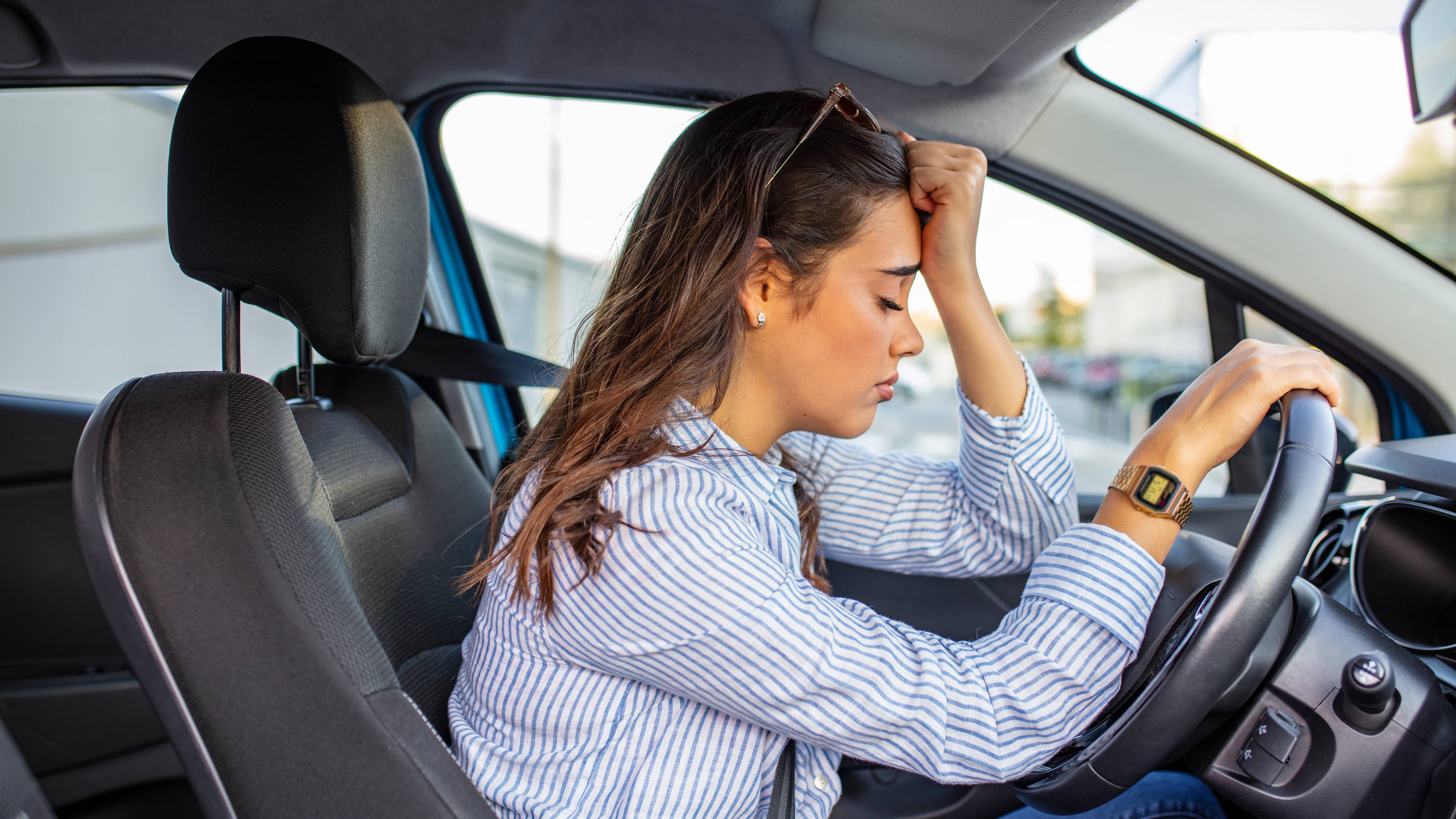 Fahrangst Header 16:9 Zu sehen ist eine Frau, die angegurtet in einem stehenden Auto sitzt. Sie lehnt gegen das Lenkrad und hält sich mit der linken Hand die Stirn.