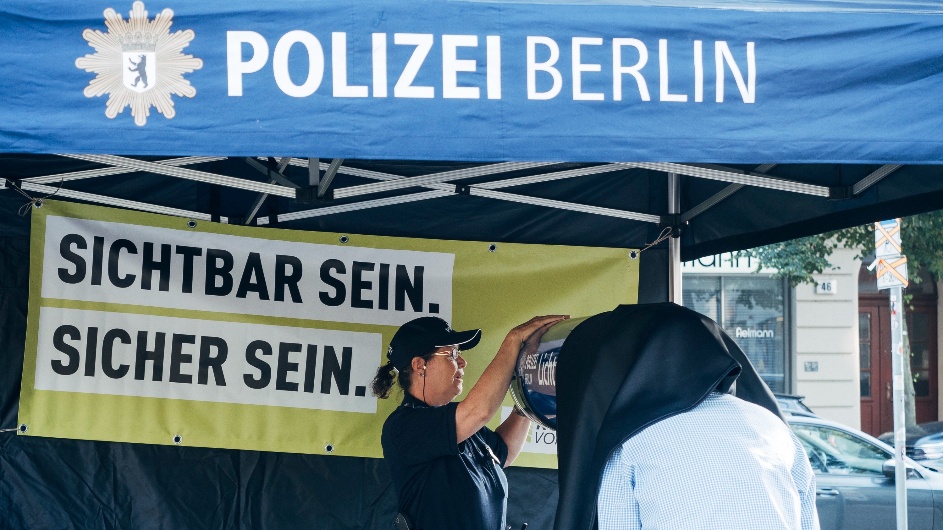 Ein Mann mit einem hellblauen Hemd blickt in eine Röhre, deren Öffnung mit einem Tuch abgehängt ist. Es ist der Lichttunnel der Polizei Berlin. 