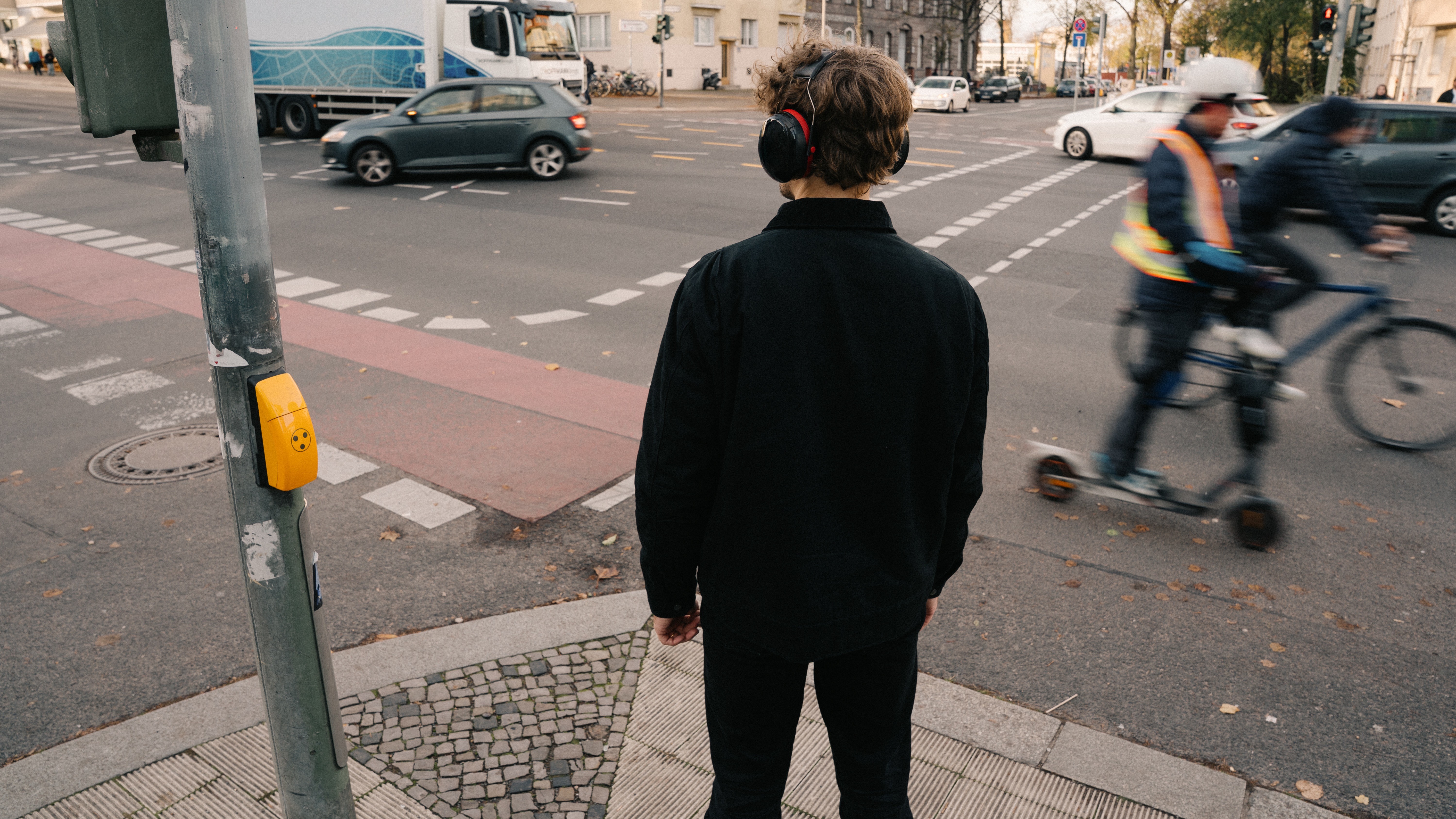 Eine Person mit Gehörschutz wartet an einer belebten Straßenkreuzung. Auf der Straße herrscht viel Verkehr mit Autos, einem Lastwagen sowie einem Radfahrer und einer Person auf einem E-Scooter.