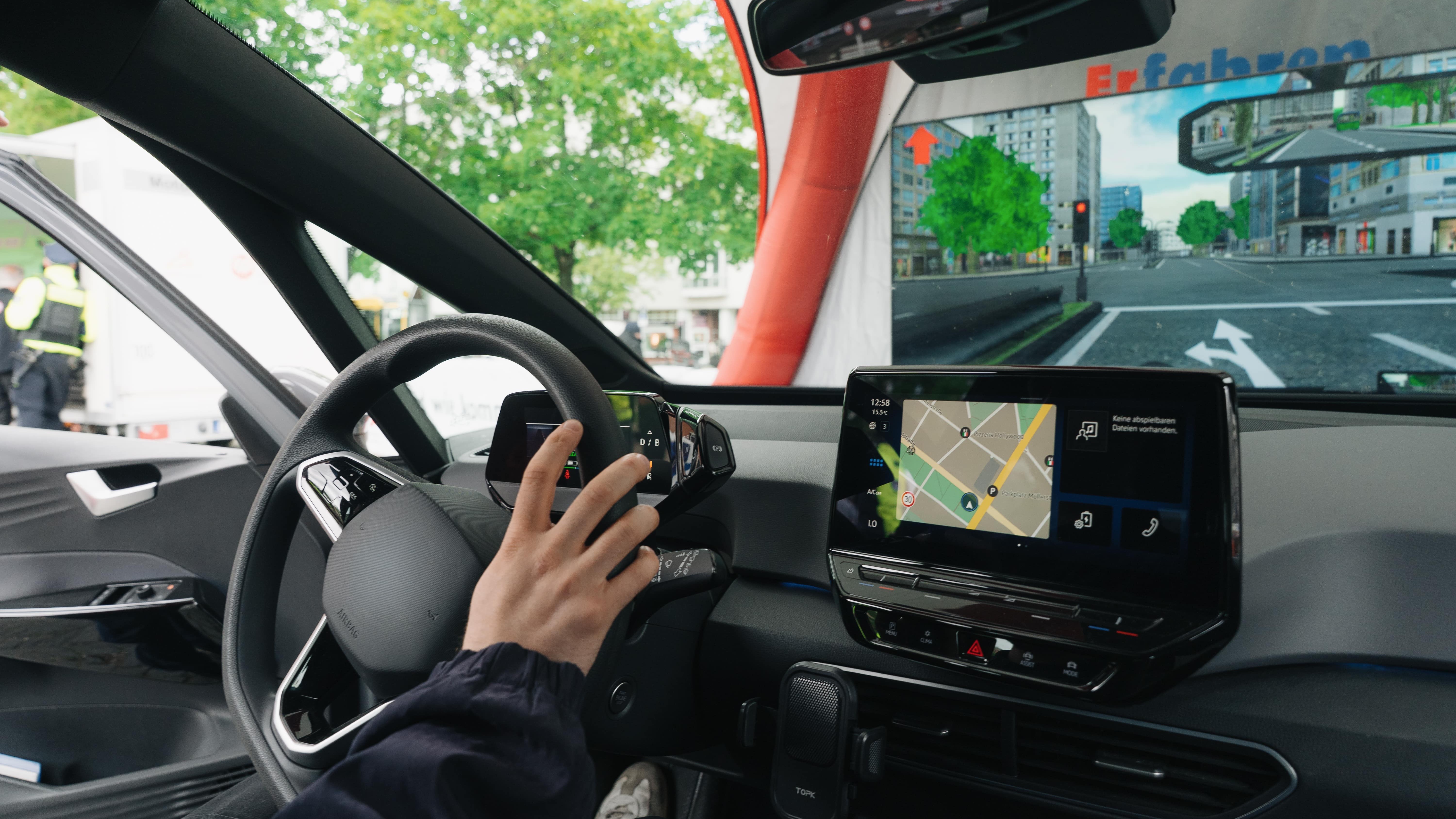 Blick aus einem Alkoholsimulator: eine Hand am Lenkrad, vor der Windschutzscheibe eine virtuelle Stadt mit Ampel und Gebäuden.