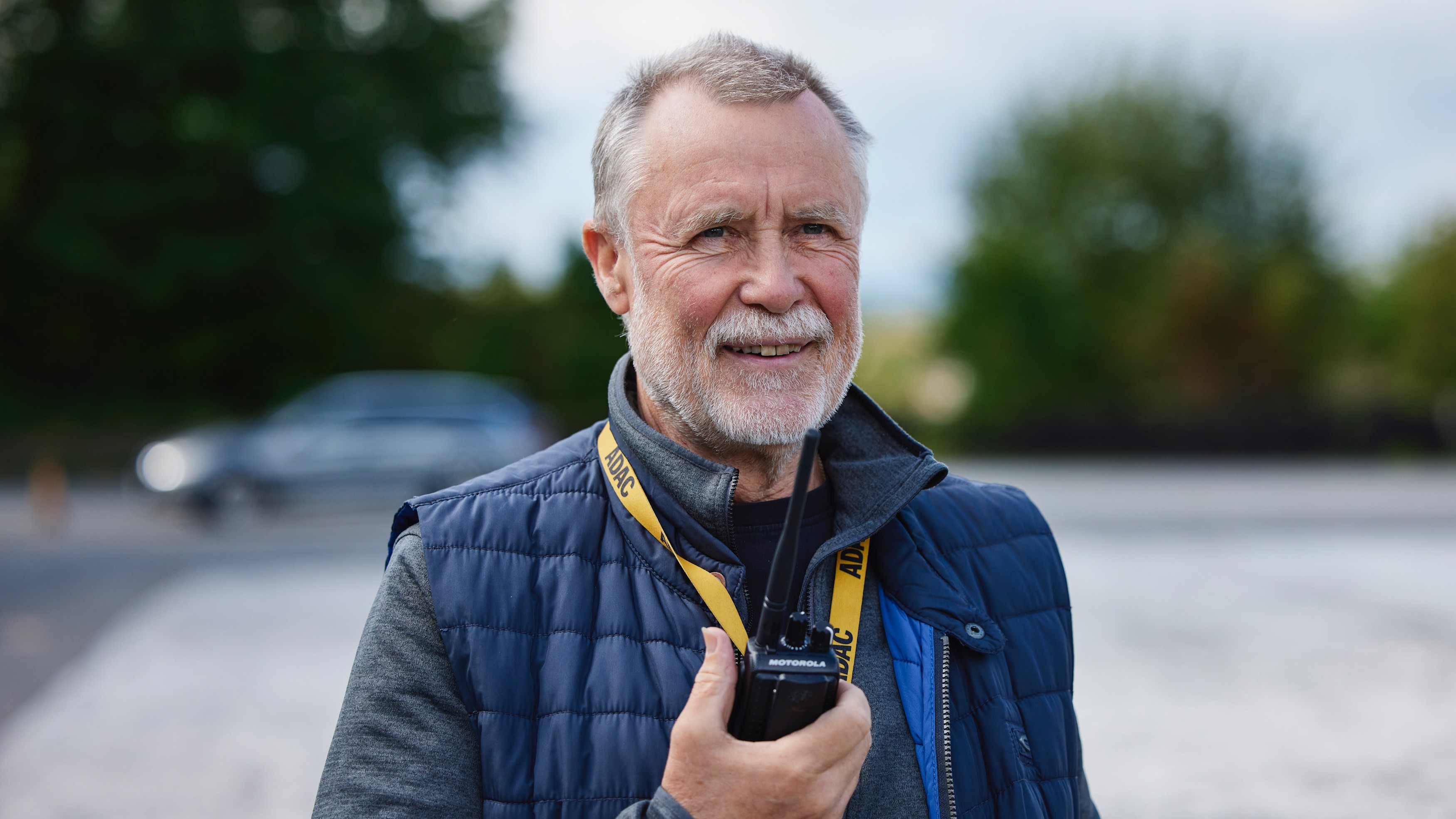 Fahrsicherheitstraining Burkhard Müller Eine Person steht im Freien und hält ein Funkgerät in der Hand. Sie trägt eine dunkelblaue Steppweste über einem grauen Pullover. Um den Hals hängt ein gelbes Schlüsselband mit der Aufschrift „ADAC". Im Hintergrund sind Bäume und ein unscharfes Auto zu sehen