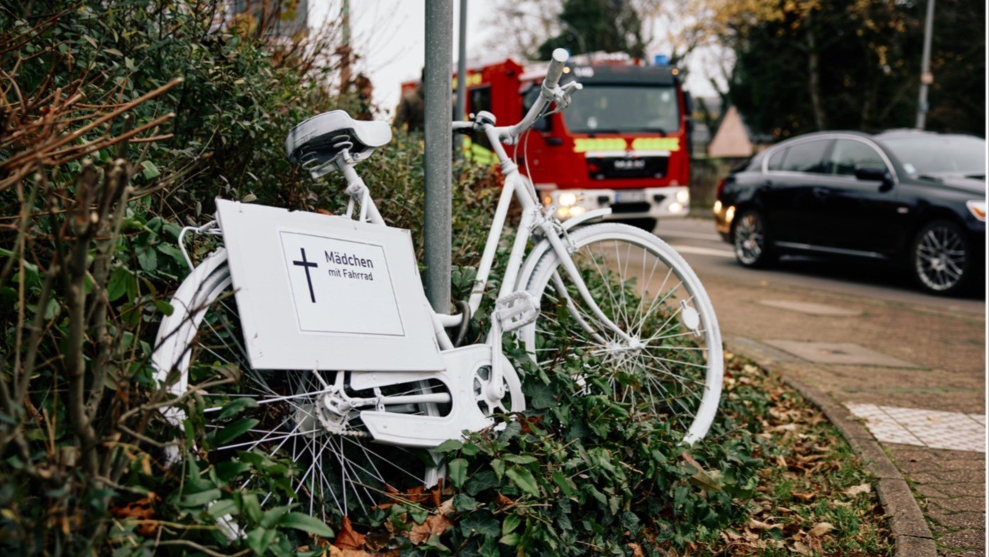 Bild weißes Rad Gedenken Ein weiß lackiertes Fahrrad steht am Straßenrand in einem Gebüsch. An dem Fahrrad ist ein Schild mit einem schwarzen Kreuz und der Aufschrift „Mädchen mit Fahrrad“ befestigt.