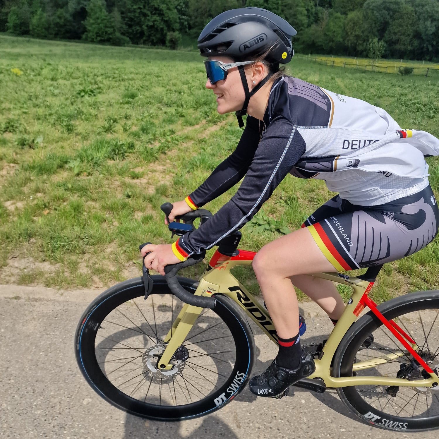 mehrAchtung Maike Hausberger Eine Frau fährt in Fahrradkleidung und Helm Rennrad auf einer Straße. Im Hintergrund sind eine Wiese und Bäume zu sehen.