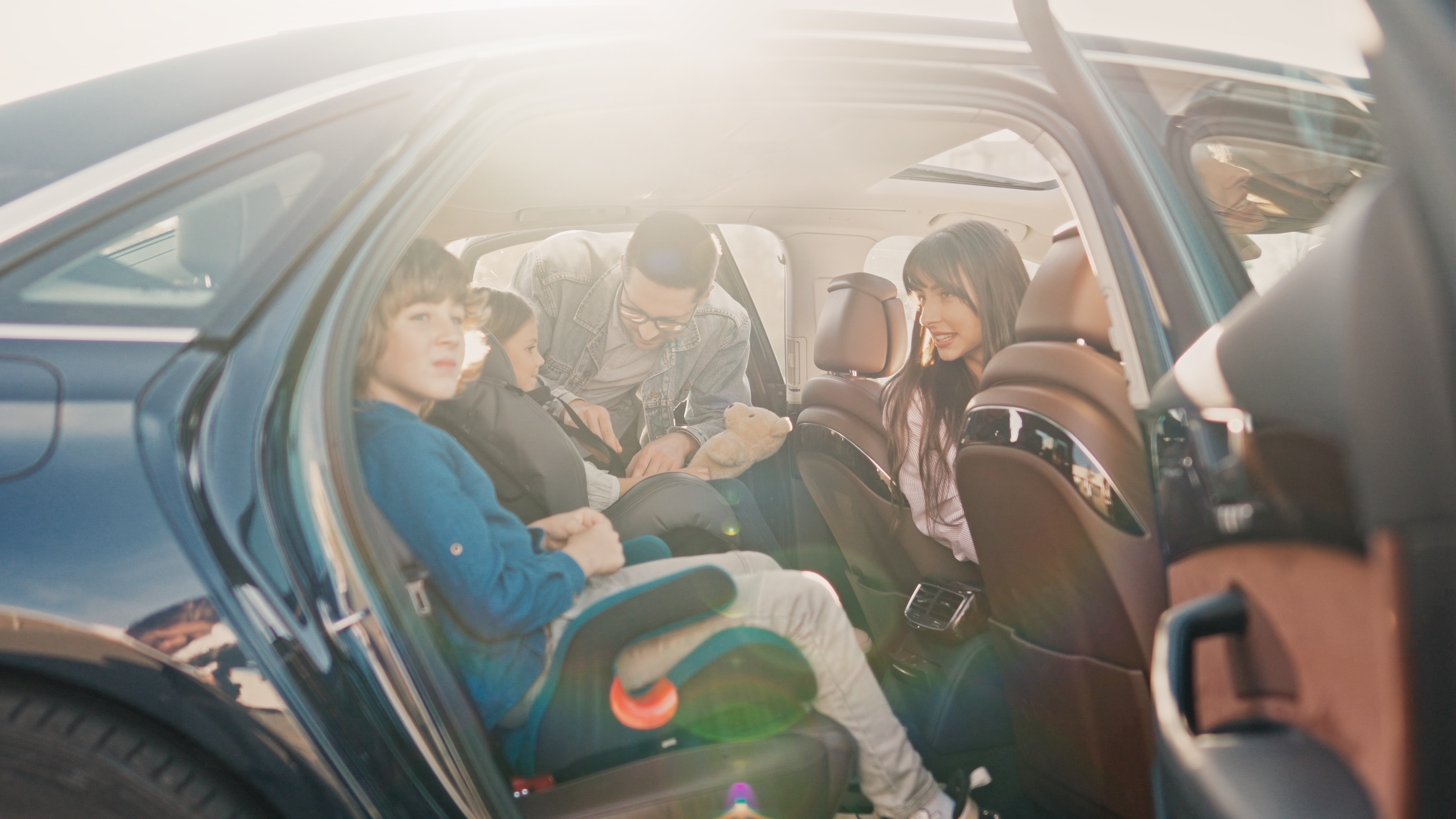 50 Jahre Anschnallpflicht mehrere Menschen im Auto Eine Familie bereitet sich auf eine Autofahrt vor. Ein Kind sitzt angeschnallt im Kindersitz auf der Rückbank, während die Eltern beim Einsteigen helfen.