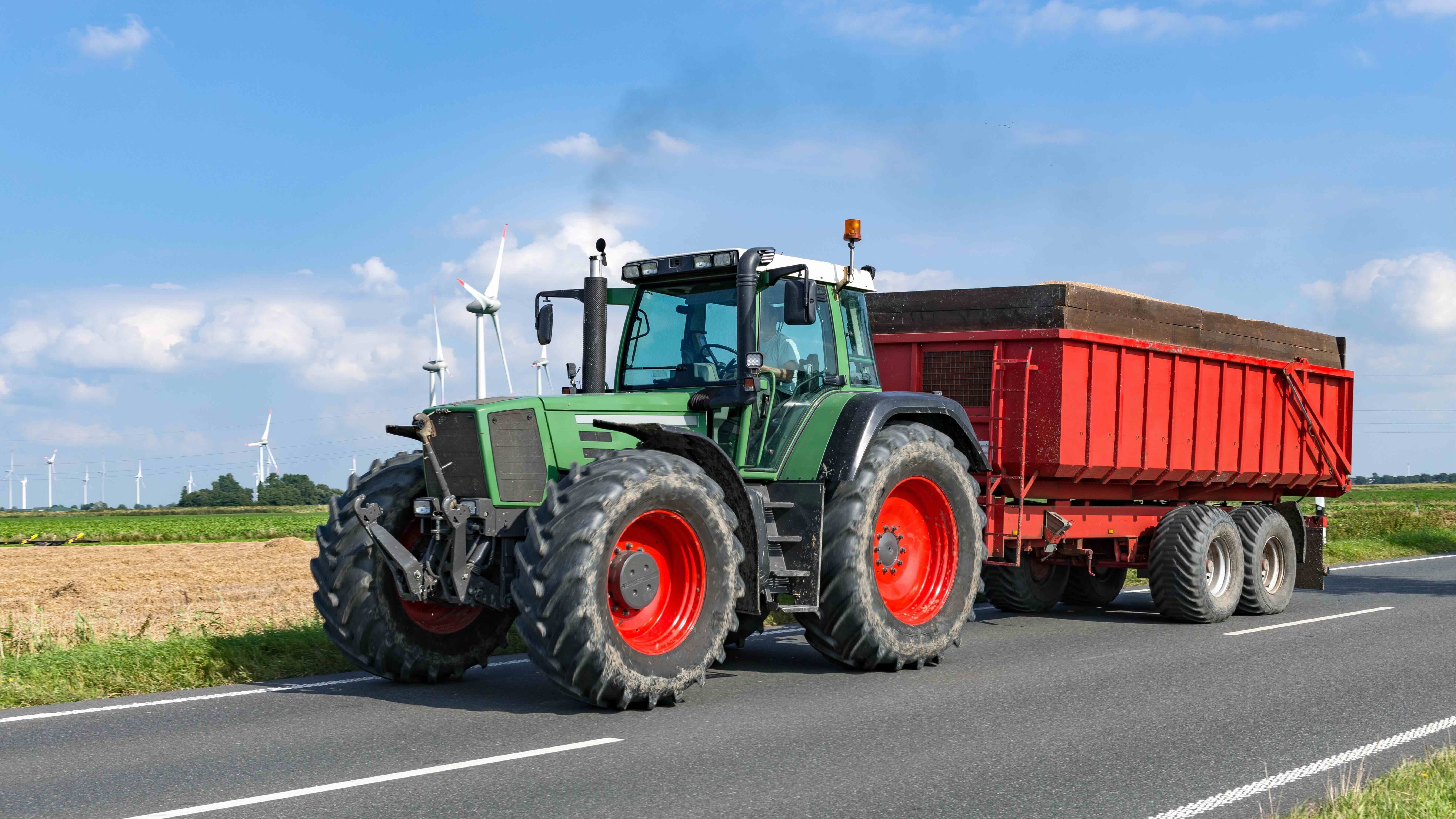 Traktor auf Landstraße Ein grüner Traktor zieht einen roten Anhänger auf einer asphaltierten Straße. Im Hintergrund sind Windkraftanlagen und Felder unter blauem Himmel zu erkennen.
