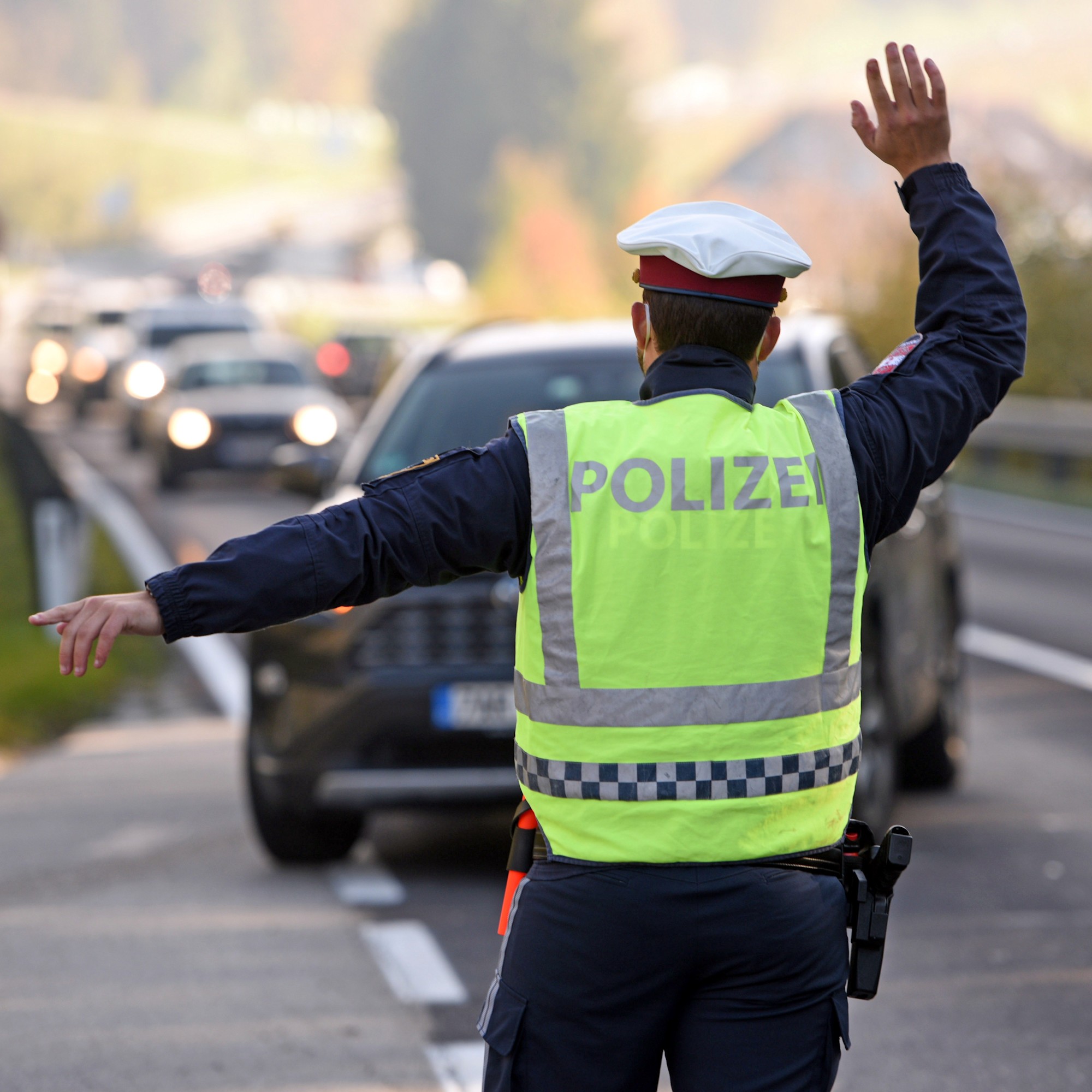 Restalkohol Polizist Das Bild zeigt einen Polizisten von hinten, der auf einer Straße steht und den Verkehr regelt. Er trägt eine dunkelblaue Uniform leuchtend gelb-grüne Warnweste mit der Aufschrift "POLIZEI" auf dem Rücken. Seine Arme sind ausgestreckt, wobei die rechte Hand erhoben ist, um den Verkehr anzuhalten, und die linke Hand nach links zeigt. Im Hintergrund sind mehrere Pkw zu sehen, die auf der Straße warten.