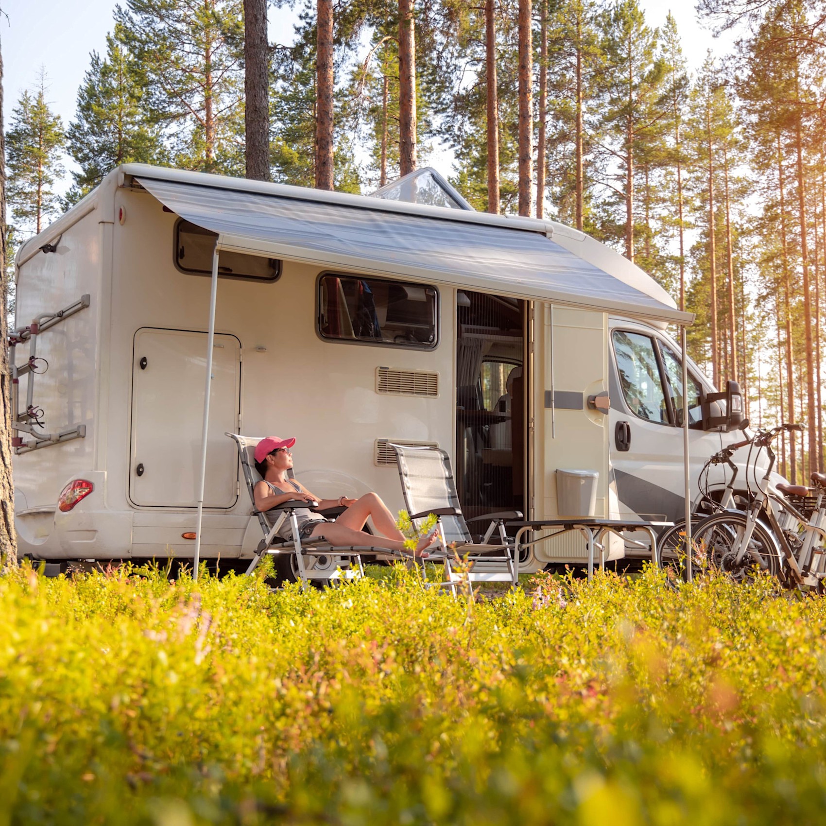 Wohnmobilurlaub – Wohnmobil im Wald Ein weißes Wohnmobil steht mit ausgefahrener Markise in einem sonnigen Wald. Vor dem Wohnmobil entspannt eine Person mit rosa Kappe in einem Liegestuhl. Fahrräder lehnen am Fahrzeug. Das Bild vermittelt die entspannte Atmosphäre eines Urlaubs mit dem Wohnmobil in der Natur und soll auf Sicherheitstipps für Wohnmobilreisende verweisen.