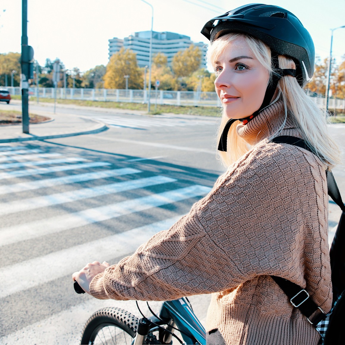 Eine blonde Frau schiebt ihr Fahrrad lächelnd über den Zebrastreifen im Stadtgebiet.