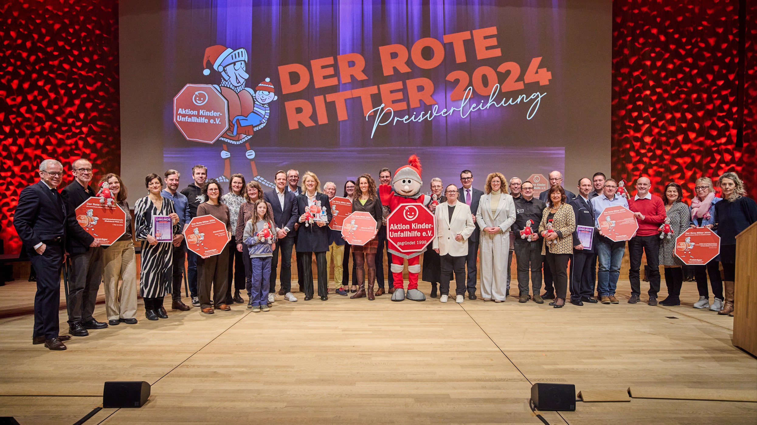 16:9 Header Roter Ritter Auf einer Bühne steht eine Gruppe von Frauen, Männern und einem Kind. In der Mitte steht ein großes Ritter-Maskottchen. Alle lächeln in die Kamera. Es sind die Vorsitzenden der Aktion Kinder-Unfallhilfe e. V. und die Preisträgerinnen und Preisträger von „Der Rote Ritter“ 2024.