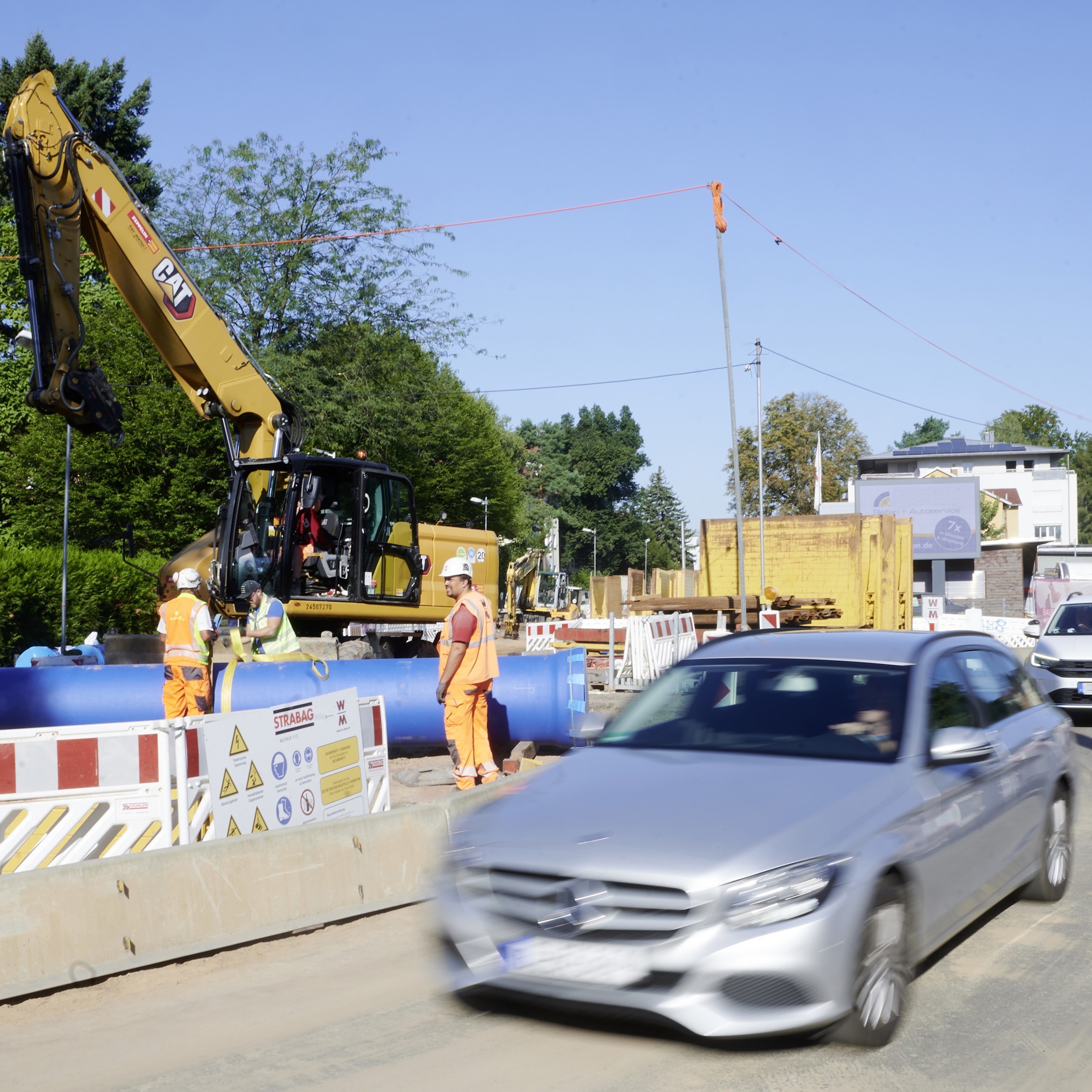 Eine Baustelle auf einer Straße mit laufendem Verkehr. Im Hintergrund sind Bauarbeiter, ein Bagger und große blaue Rohre zu sehen. Im Vordergrund fährt ein silbernes Auto vorbei. Die Baustelle ist mit rot-weiß gestreiften Absperrungen gesichert.
