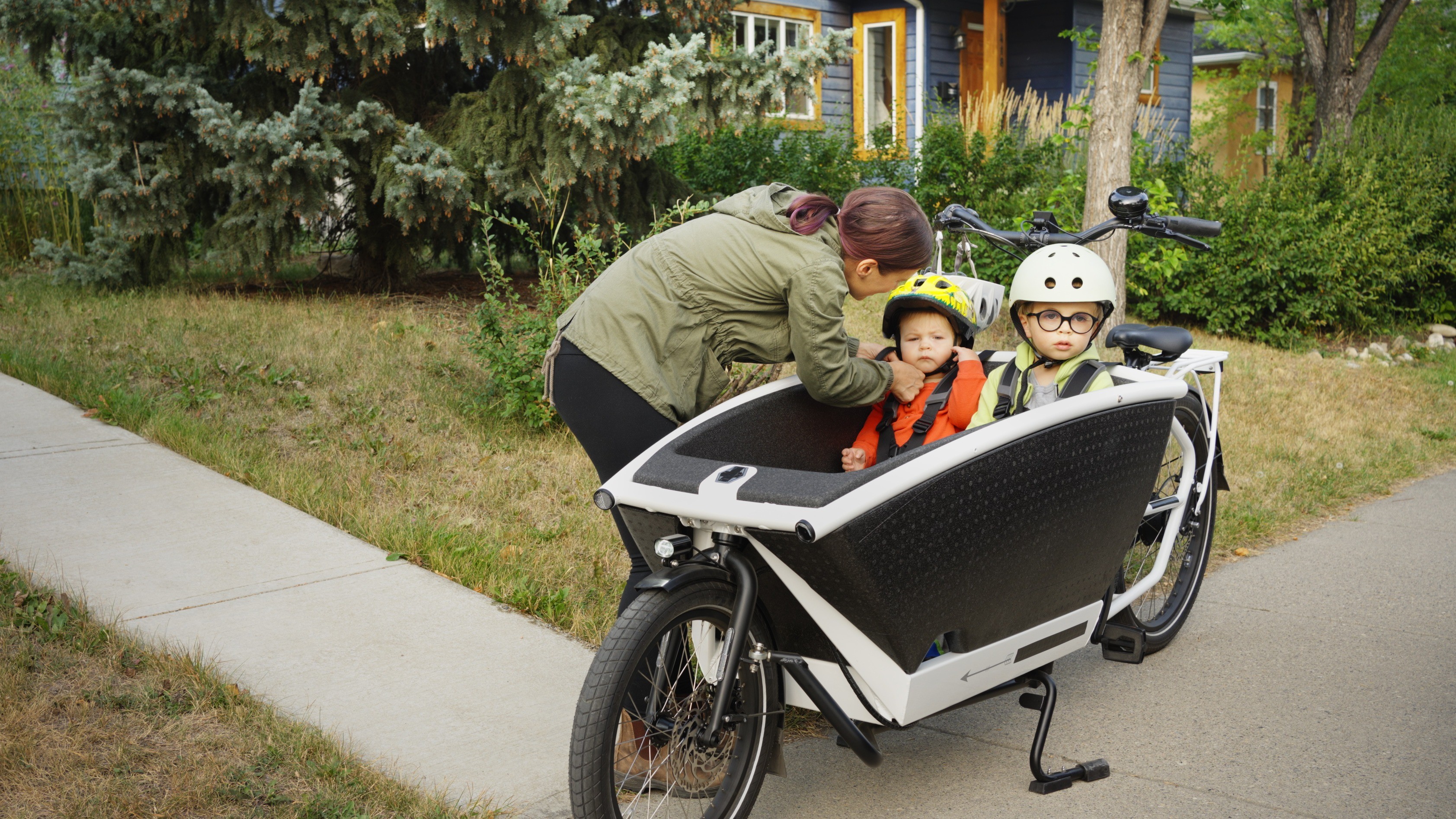 Lastenrad Kinder Zwei Kinder sitzen angeschnallt und mit Fahrradhelmen in der Box eines modernen E-Lastenrads. Eine Frau sichert die Kinder und prüft den Sitz eines Helms.