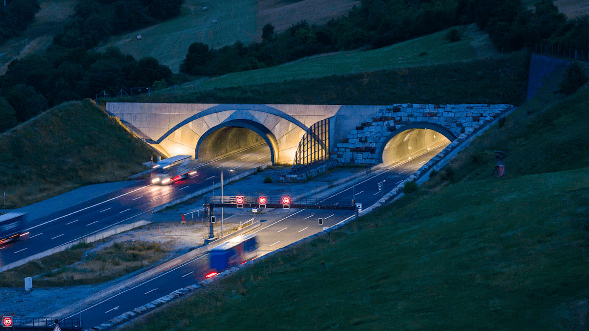 Zwei beleuchtete Straßentunnel auf einer Autobahn bei Dämmerung. Lkw fahren in die Röhren hinein und wieder heraus. Vor einem Tunneleingang sind rot leuchtende Verkehrsschilder zu sehen. Die Umgebung ist eine grüne Hügellandschaft.