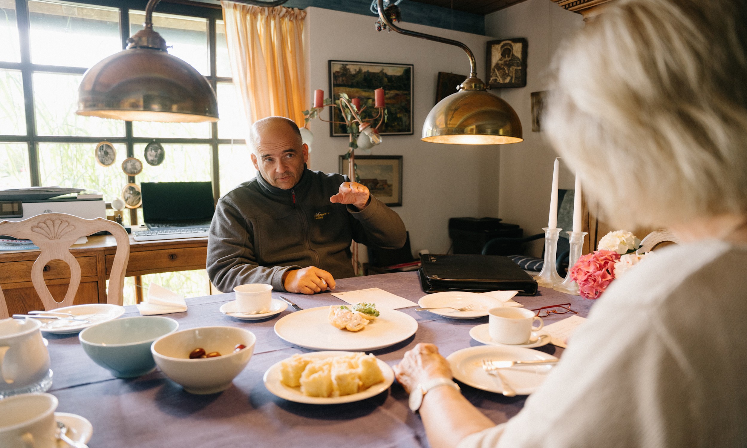 Rueckmeldefahrten Esstisch Ein Mann in einer grauen Jacke und eine ältere Frau sitzen sich an einem Esstisch gegenüber und führen ein Gespräch. Auf dem Tisch stehen Kaffeetassen, Teller mit Kuchen und ein Laptop im Hintergrund.
