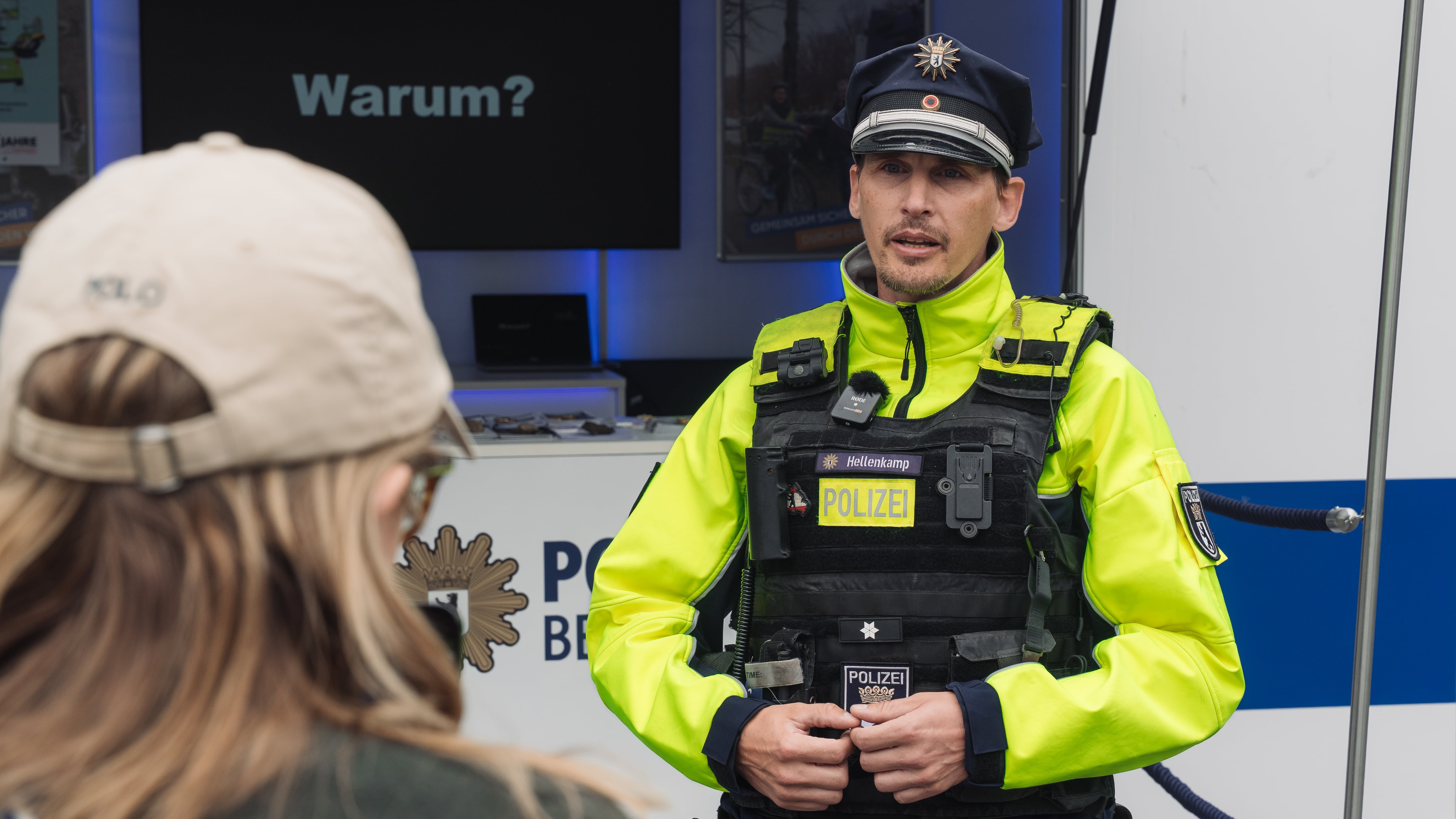 Polizeibeamter Markus Hellenkamp steht in gelber Einsatzjacke vor einem Infostand der Polizei Berlin zur Verkehrssicherheitsprävention. Im Vordergrund eine Besuchsperson mit Basecap – von hinten aufgenommen. 