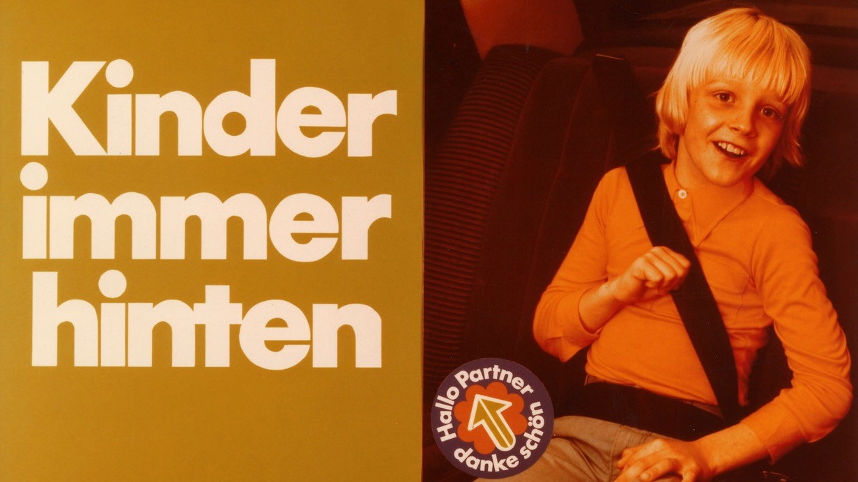 Anschnallpflicht Kinder immer hinten Lächelndes Kind sitzt im Auto angeschnallt; daneben steht der Slogan „Kinder immer hinten“.