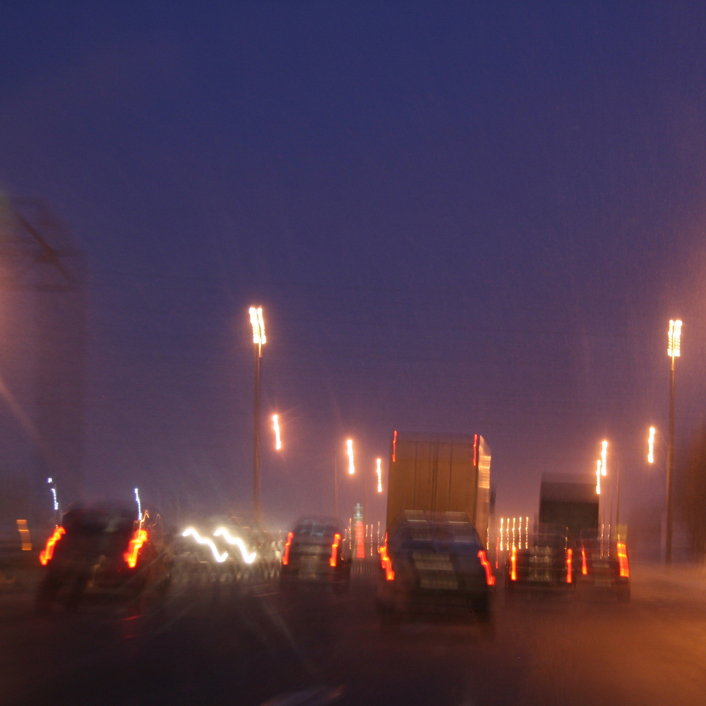Unscharfe Sicht aus einem fahrenden Auto bei Nacht. Die Lichter der Fahrzeuge und Straßenlaternen sind verschwommen und zu hellen Streifen verzogen, was eine gefährliche Verkehrssituation andeutet.