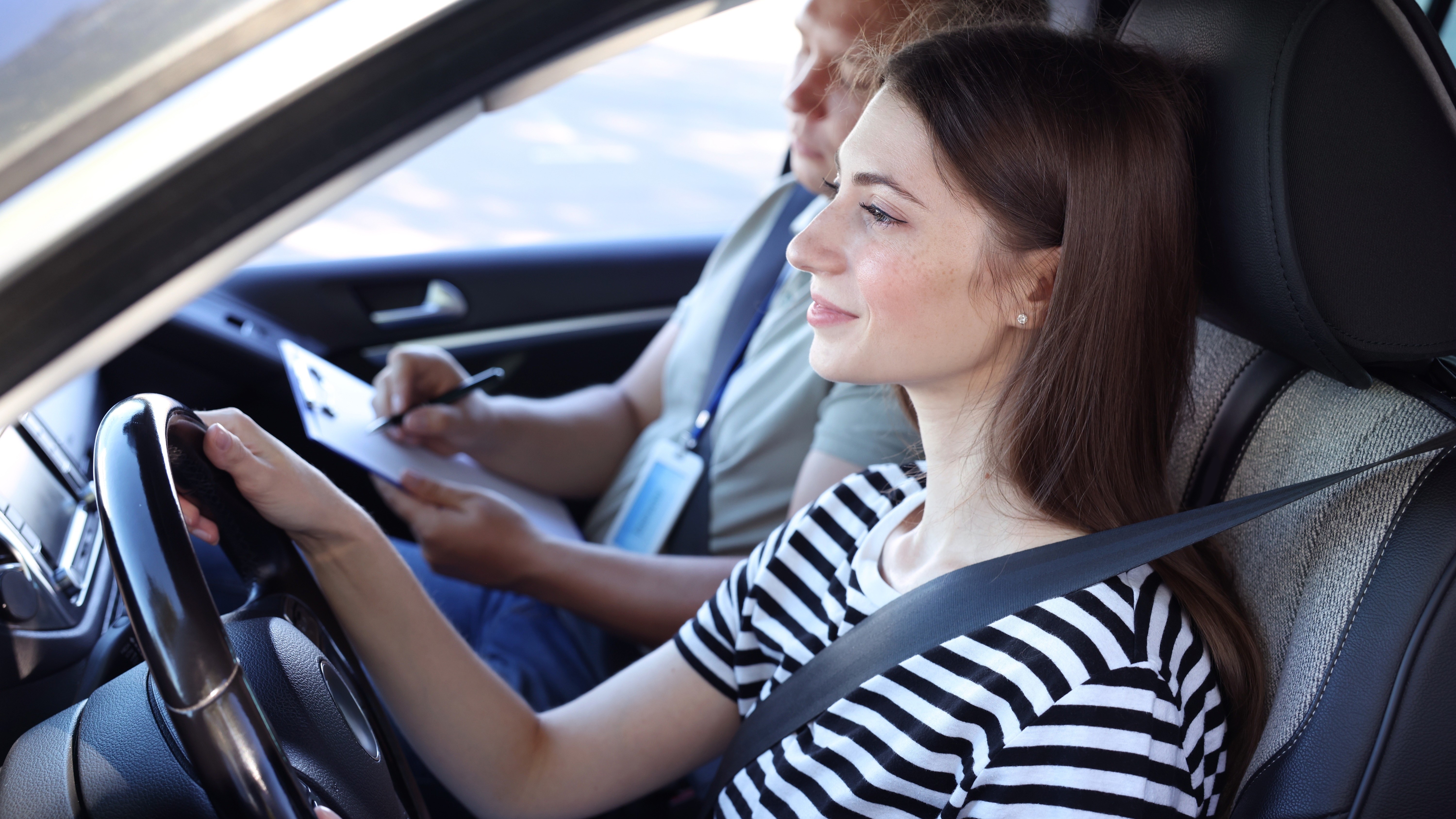 Führerscheinprüfung – Fahrstunde Eine junge Frau mit braunen Haaren sitzt auf dem Fahrersitz eines Autos und hält das Lenkrad fest. Sie trägt ein schwarz-weiß gestreiftes T-Shirt und ist mit einem Sicherheitsgurt angeschnallt. Ein Mann sitzt auf dem Beifahrersitz und macht sich Notizen auf einem Klemmbrett.