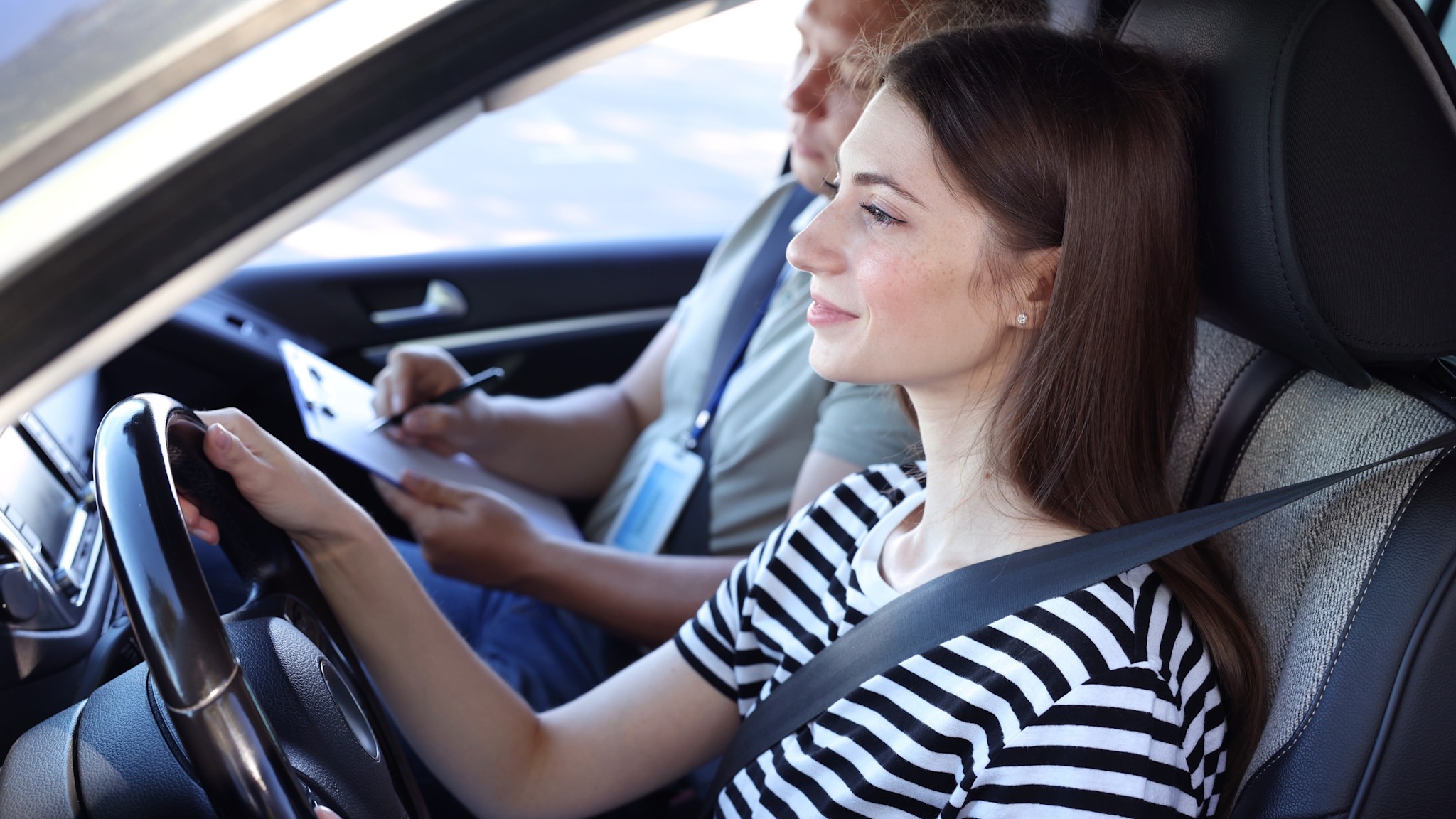 Führerscheinprüfung – Fahrstunde Eine junge Frau mit braunen Haaren sitzt auf dem Fahrersitz eines Autos und hält das Lenkrad fest. Sie trägt ein schwarz-weiß gestreiftes T-Shirt und ist mit einem Sicherheitsgurt angeschnallt. Ein Mann sitzt auf dem Beifahrersitz und macht sich Notizen auf einem Klemmbrett.