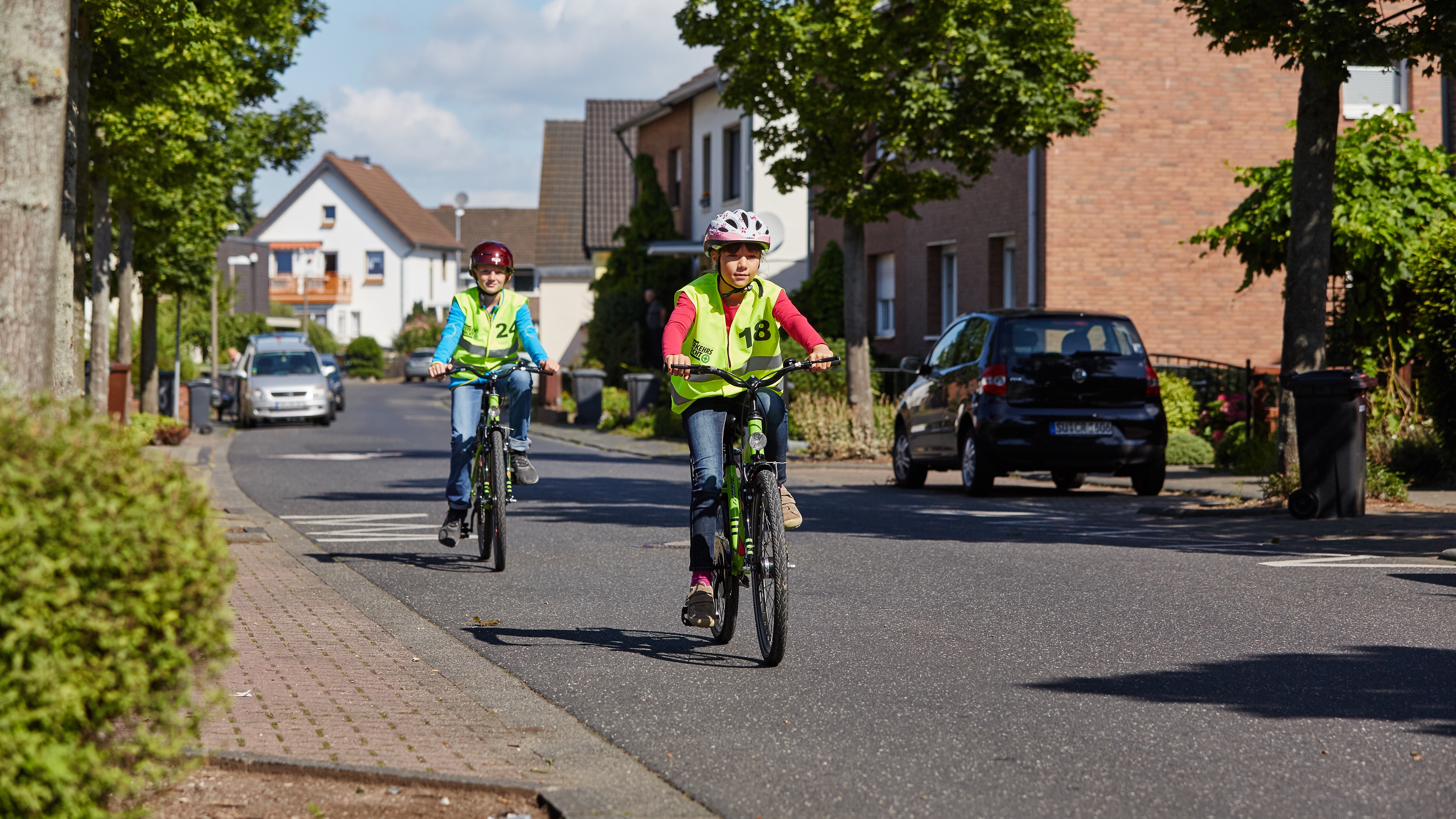 Zwei Kinder mit Helm auf Fahrrad 16:9 Zwei Kinder mit Helm und Sicherheitsweste fahren mit dem Fahrrad sicher durch eine Wohnstraße.