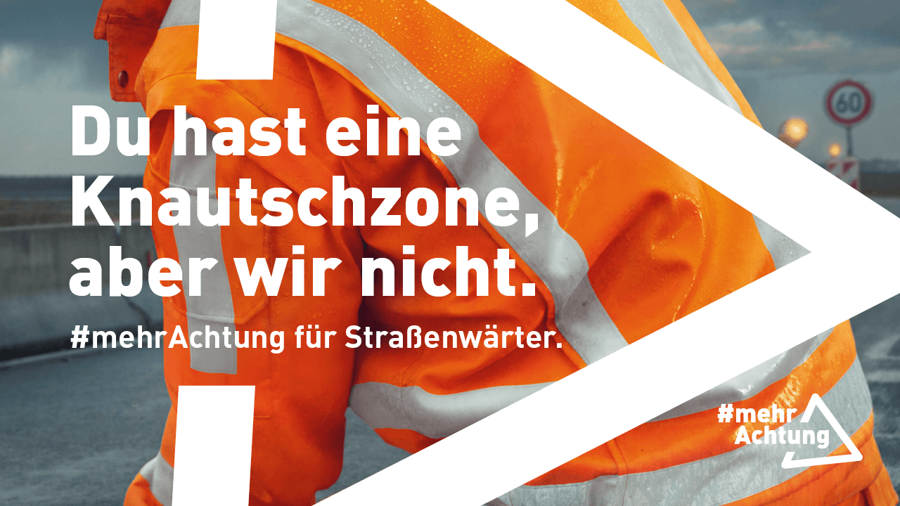 Rückansicht eines Straßenwärters in oranger Warnschutzkleidung bei der Arbeit auf einer nassen Fahrbahn. Im Hintergrund ist eine Baustelle zu erahnen. 