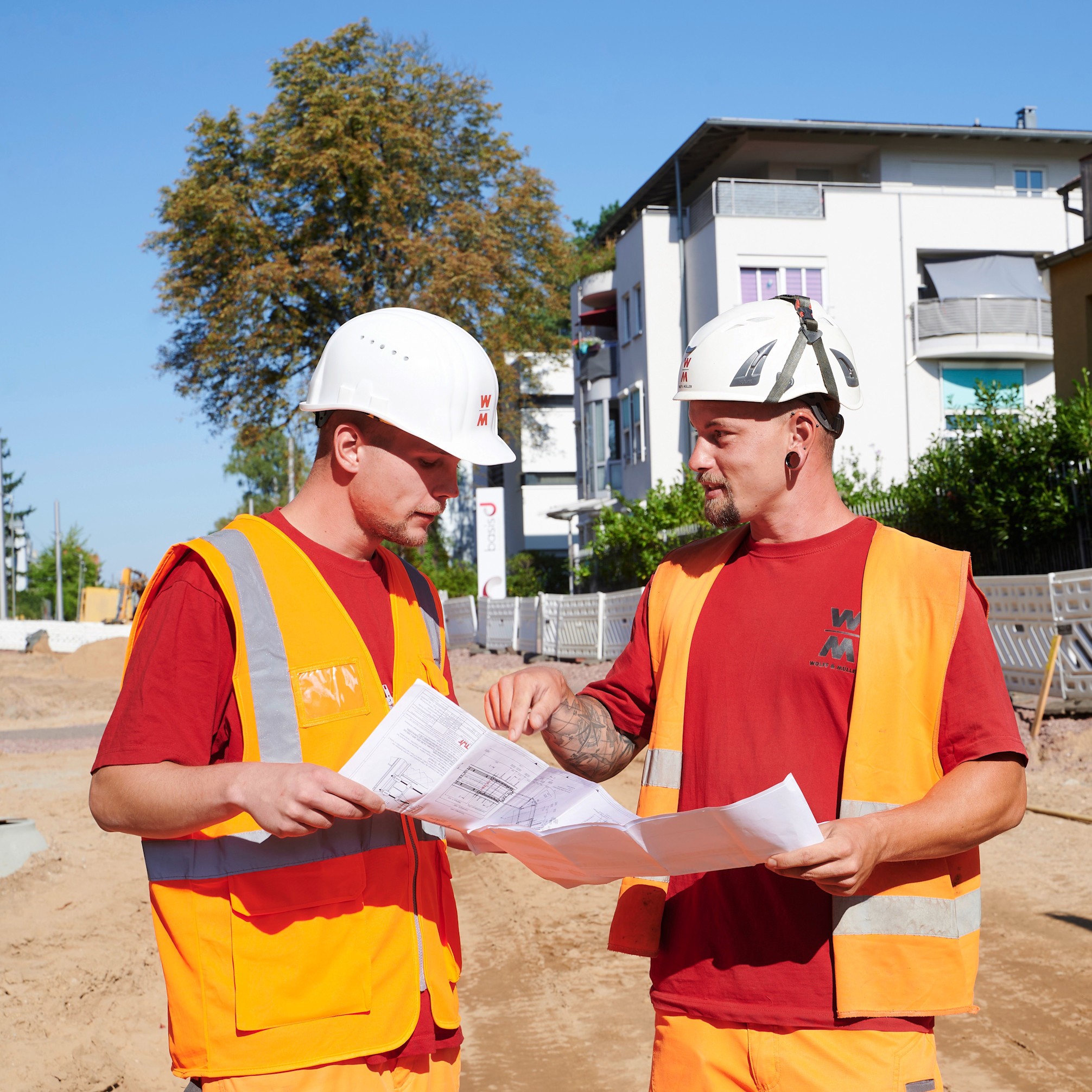 Zwei Straßenbauer in orangefarbenen Westen und Helmen betrachten gemeinsam einen Bauplan auf einer Baustelle.