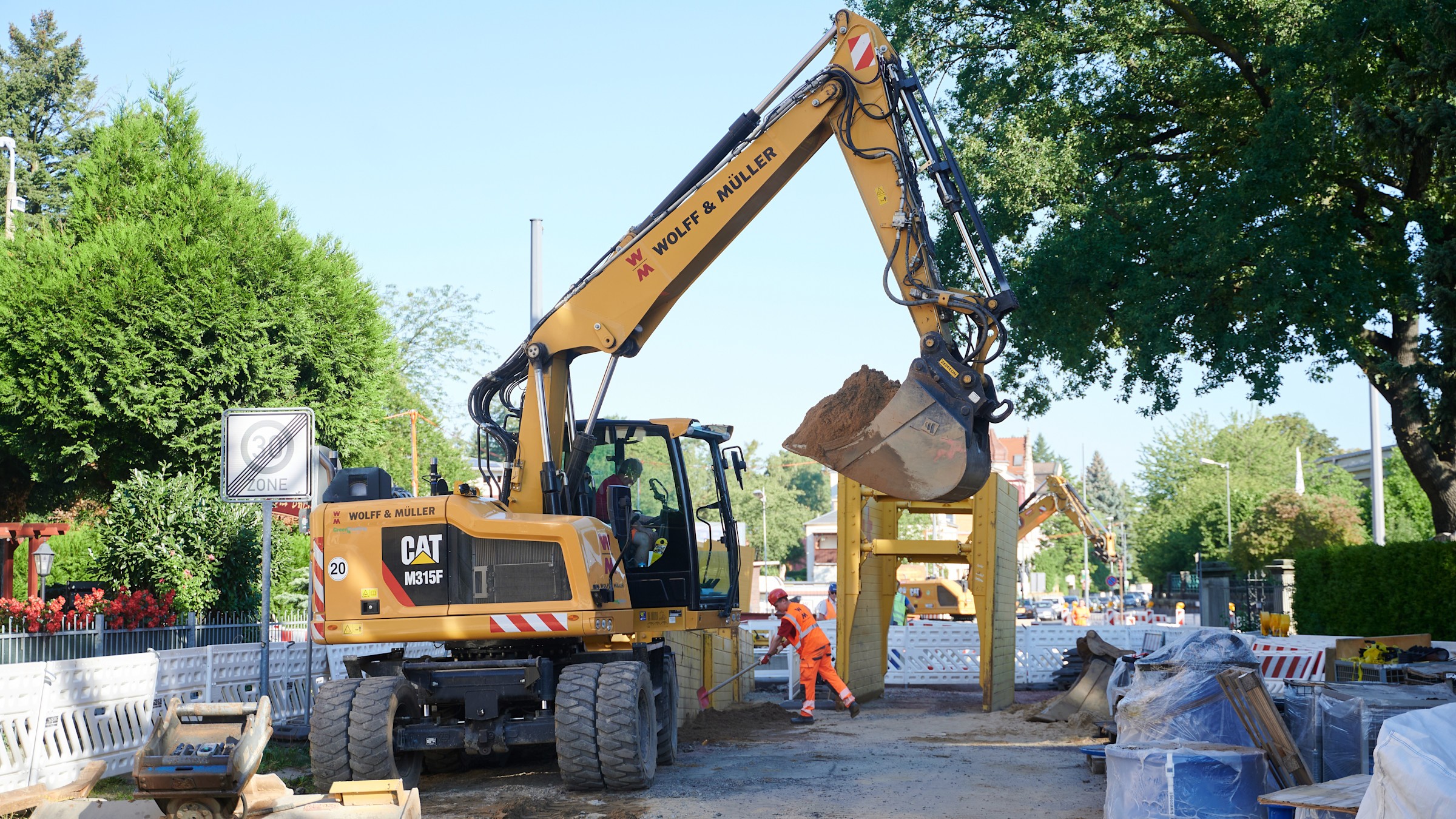 Ein gelber Bagger hebt Erde auf einer Baustelle aus. Ein Bauarbeiter in orangefarbener Warnkleidung steht in der Nähe.