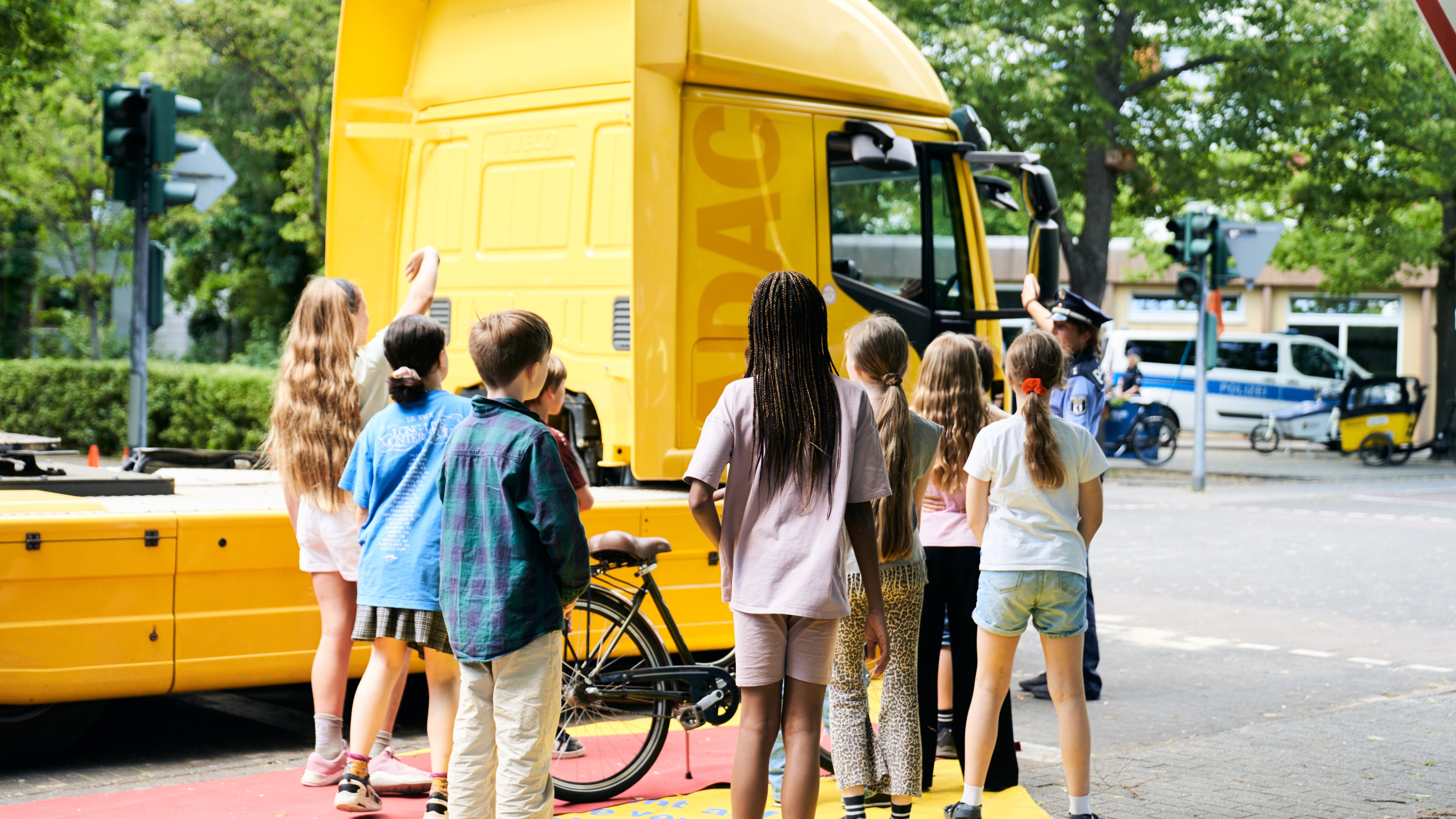 Eine Gruppe Kinder steht neben einem gelben Lkw. Auf dem Boden ist ein roter Bereich markiert, der den toten Winkel darstellt. Ein gelber Bereich daneben symbolisiert die Position, in der sie für die Person im Lkw sichtbar sind.